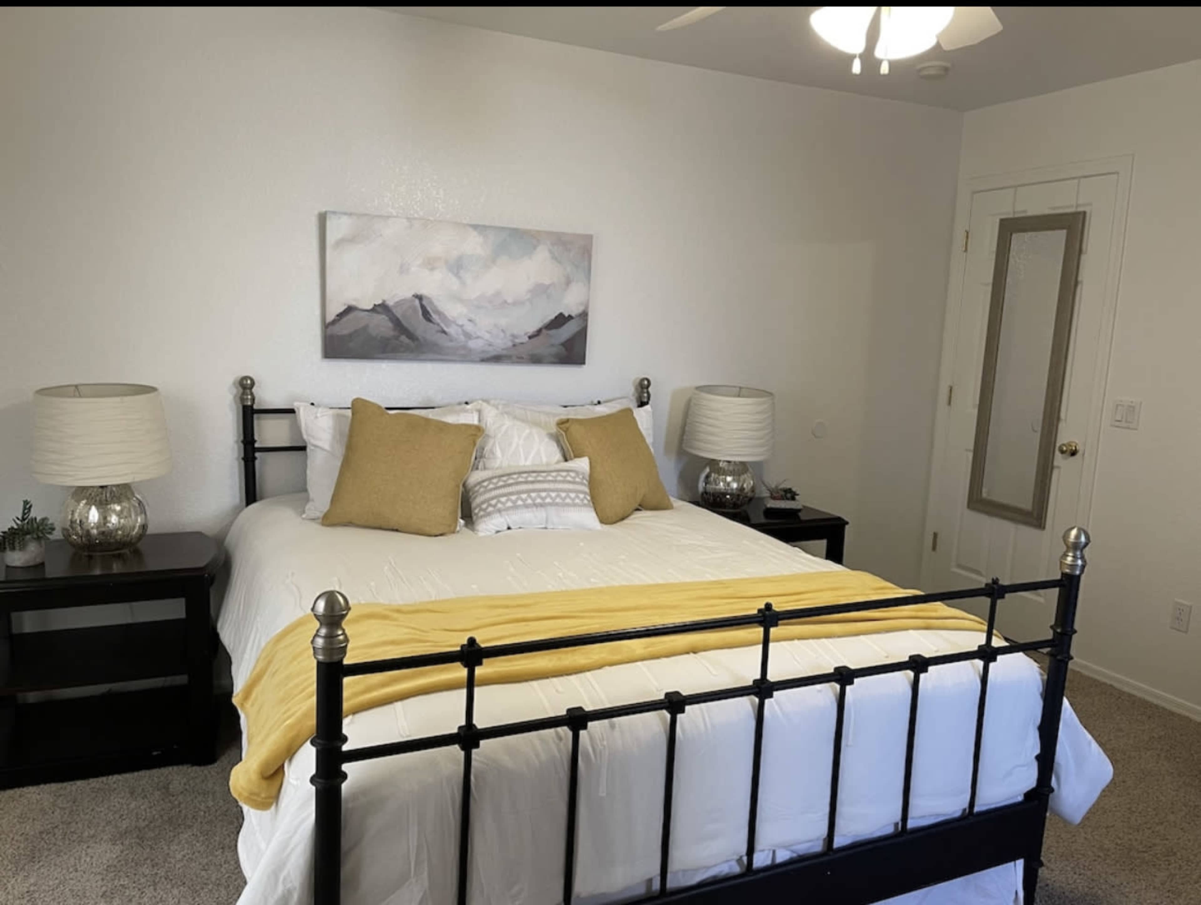 A neatly made bed with yellow and white bedding is centered between two nightstands with lamps in a well-lit bedroom.