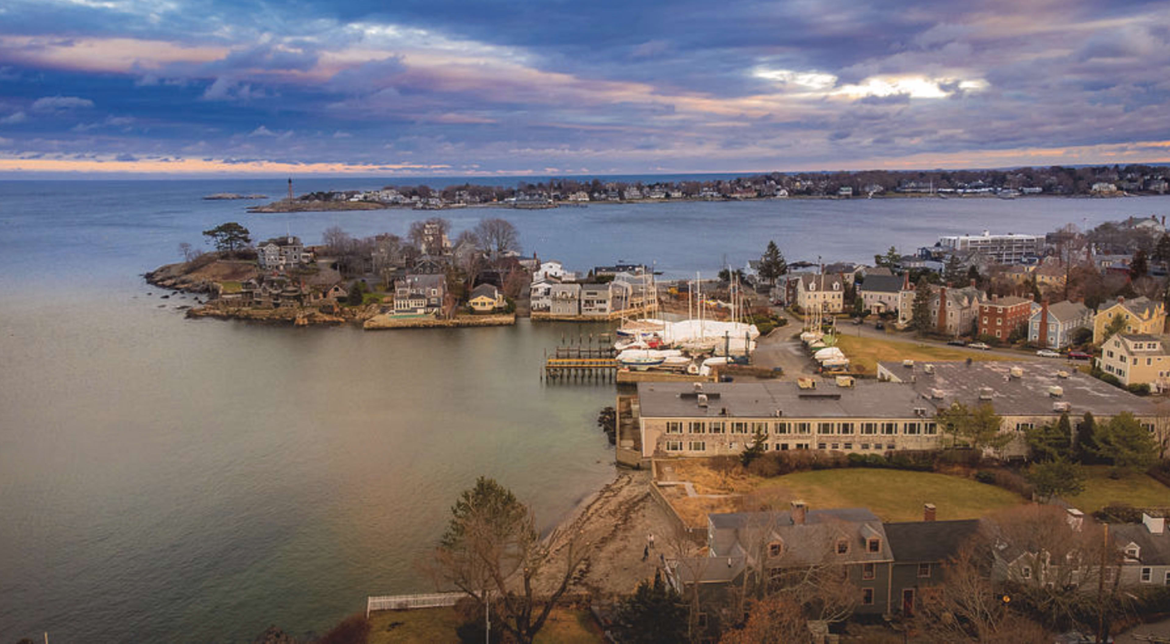 A coastal town features a marina with boats and various waterfront homes under a cloudy sky.