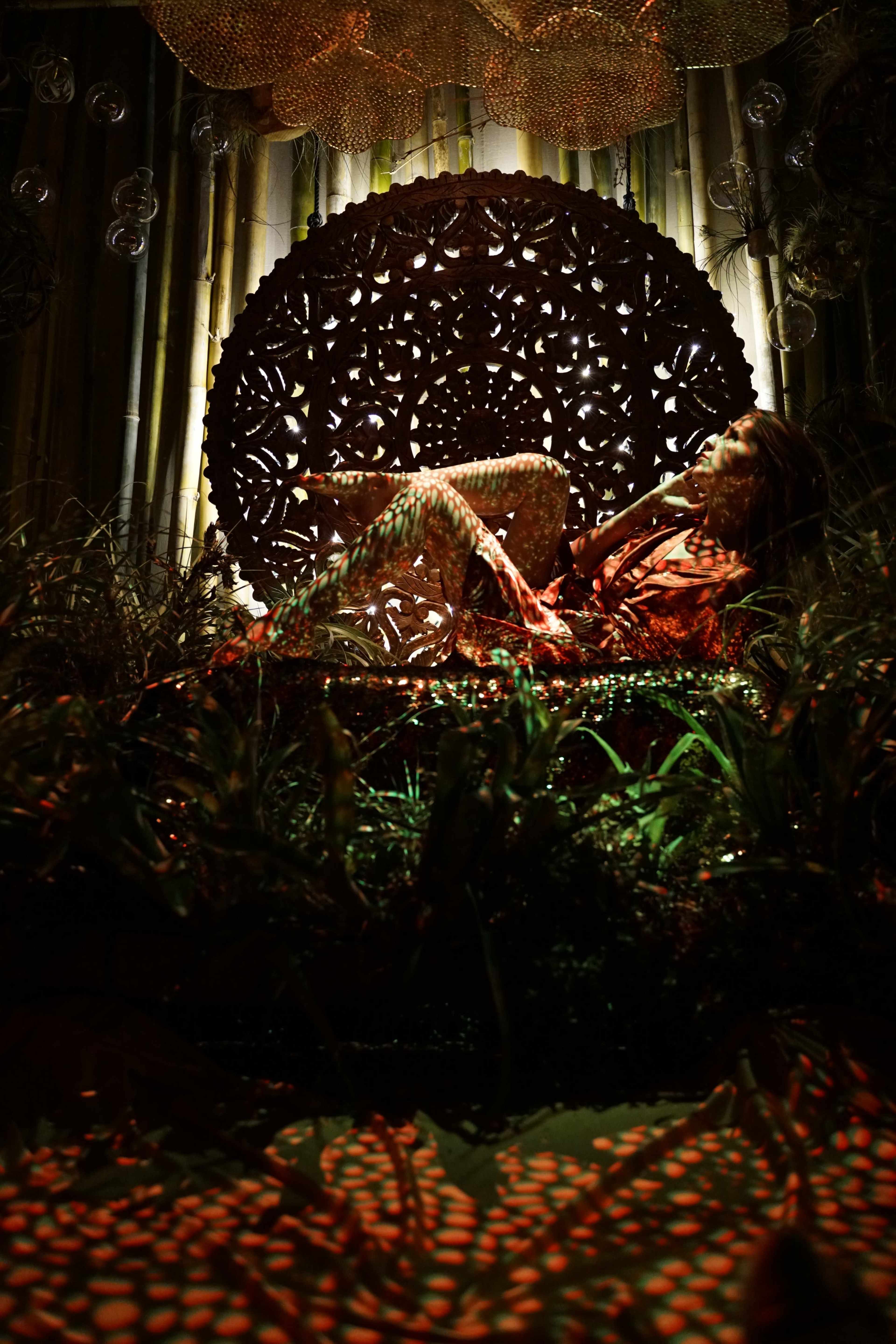 A figure poses dramatically in front of an intricate wooden backdrop, surrounded by plants and ambient lighting.