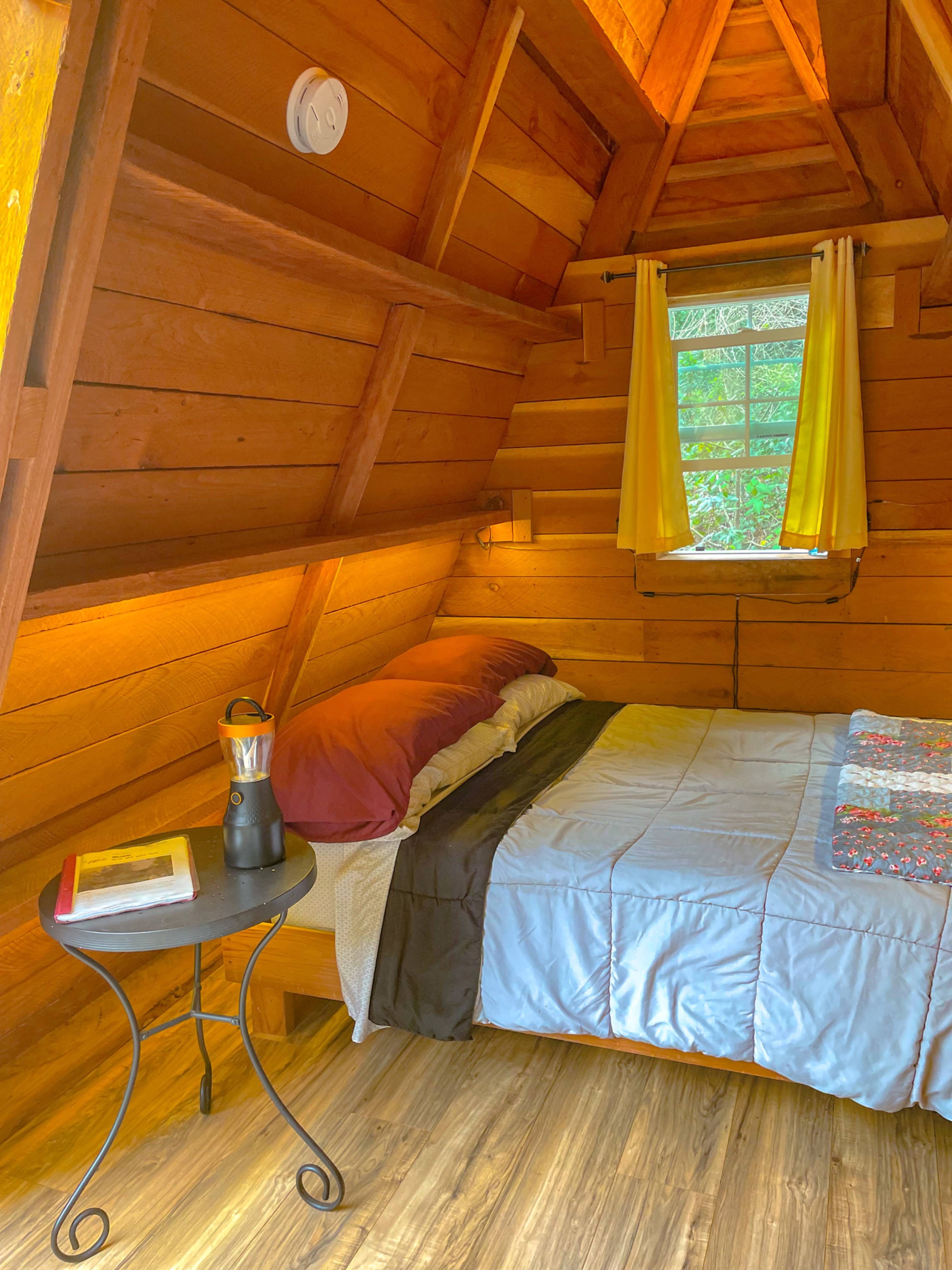 A cozy interior of a small cabin bedroom with a bed, a side table, and a window with yellow curtains, all surrounded by wooden walls.