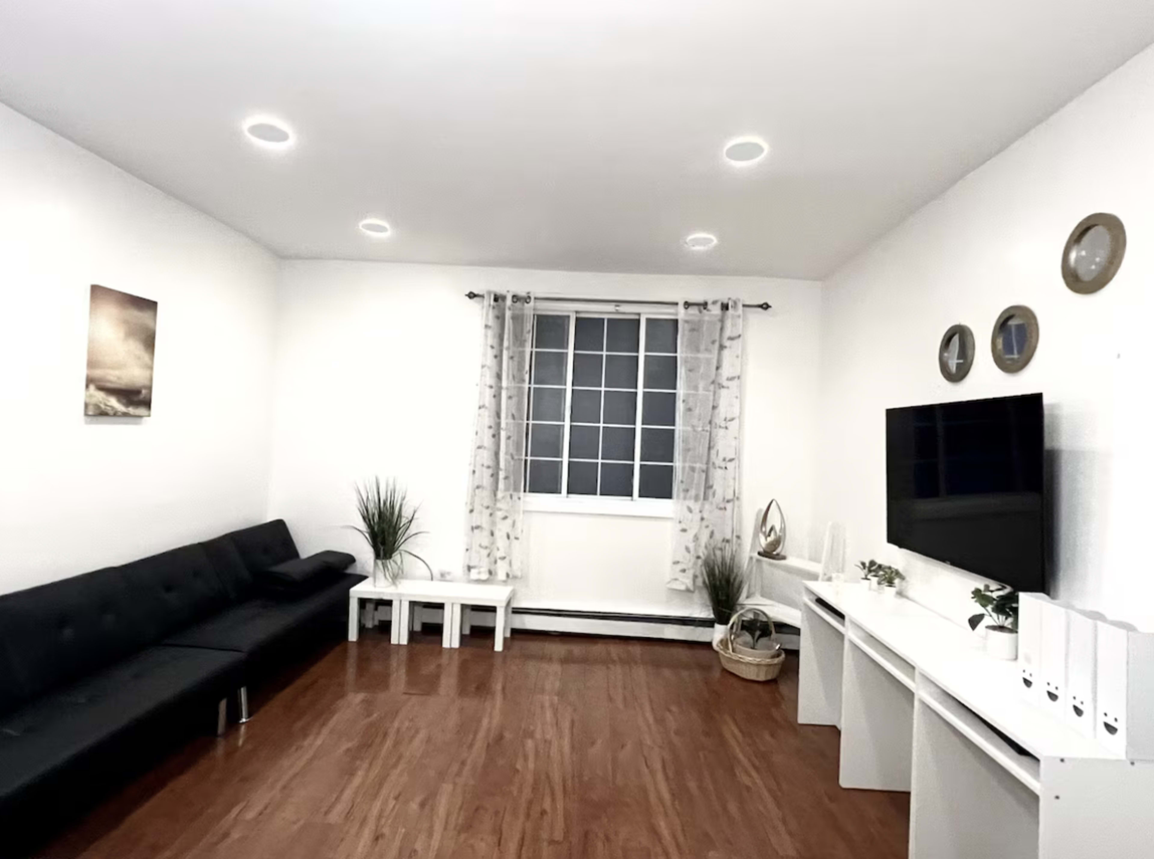 The image shows a modern living room with a black sofa, a white television console, and a window dressed with sheer curtains.