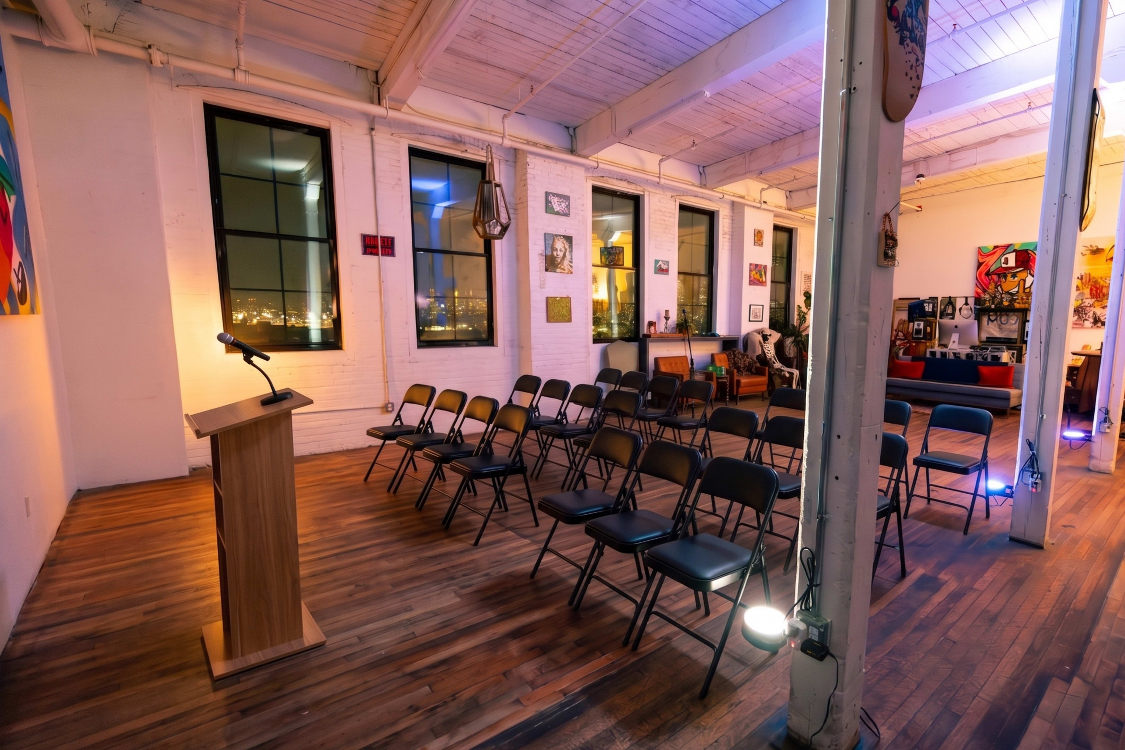 The image shows a spacious room with wooden floors, featuring a podium in front of several rows of empty black chairs, all against a backdrop of art-covered walls and large windows.