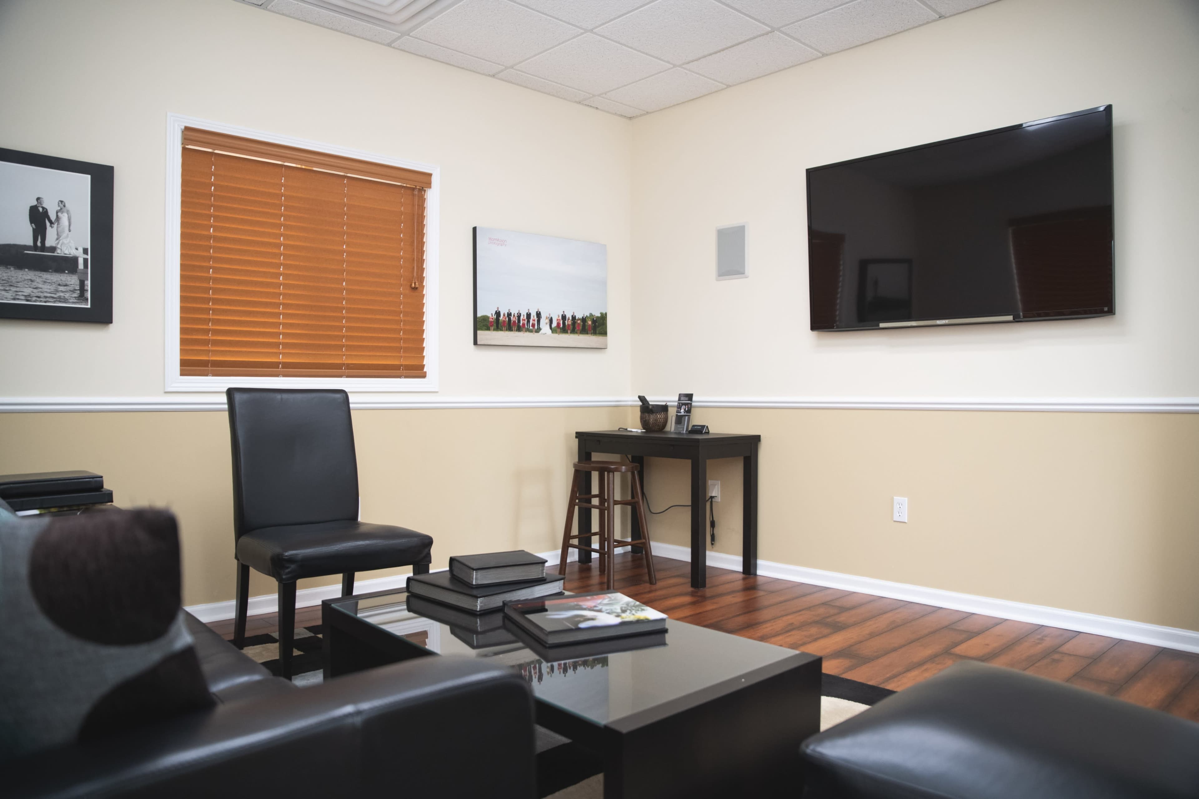 A small, modern office or meeting room with a television mounted on the wall, a black desk, a bar stool, and a few black chairs arranged around a coffee table.