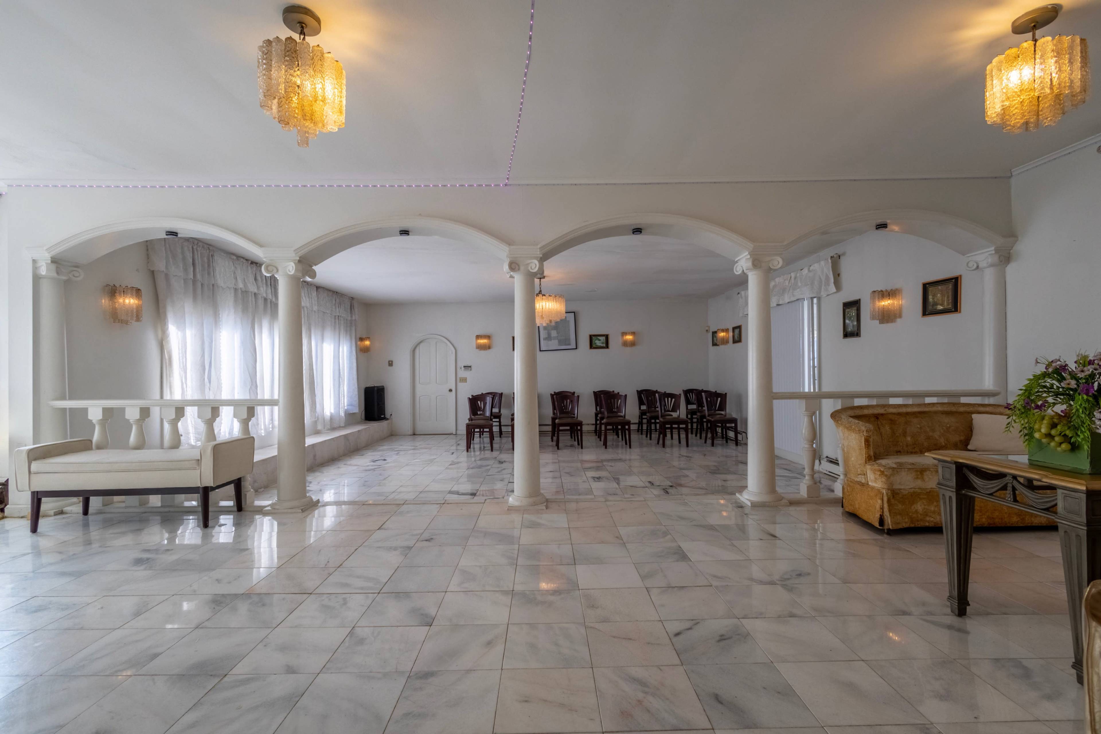 The image depicts a spacious, well-lit room with marble flooring, archways, and a seating area featuring chairs arranged around a central space.