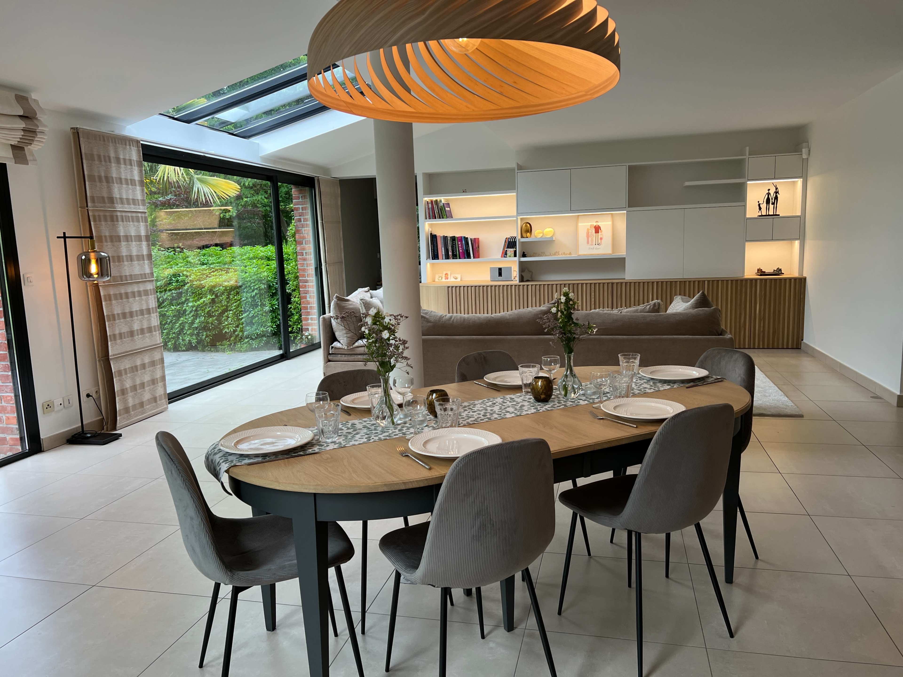 A modern dining area features a long table set for six, surrounded by gray chairs, with a large window offering a view of a garden.