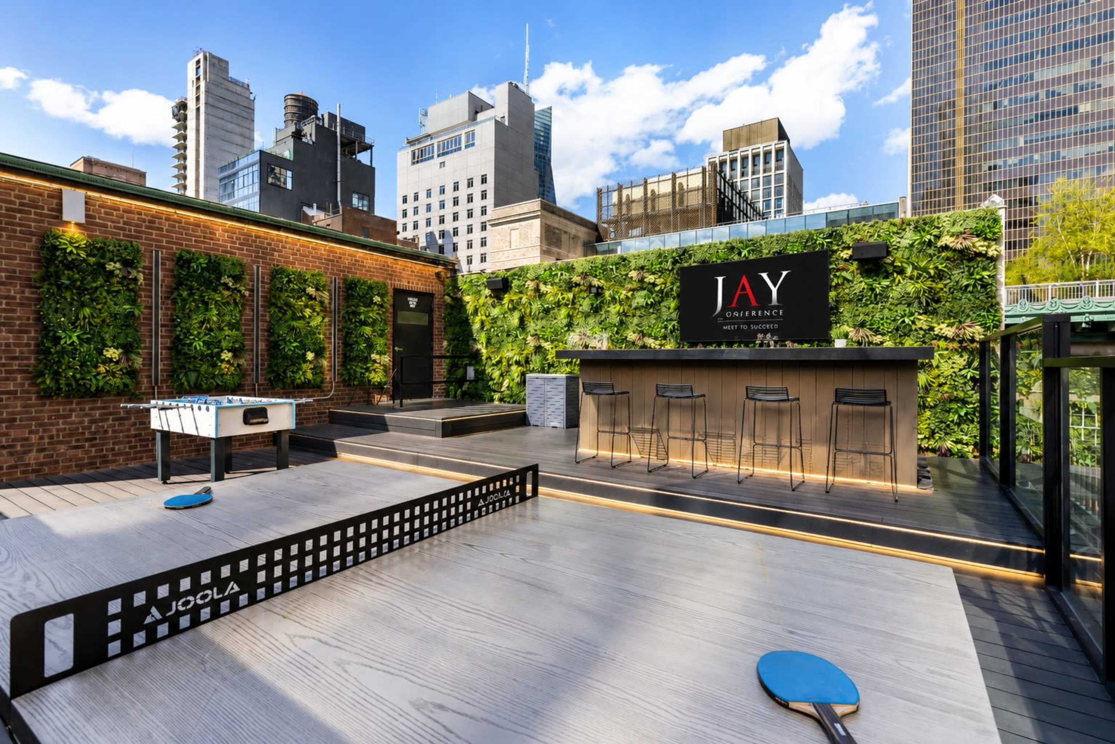 The image shows a rooftop terrace featuring a ping pong table, a bar area, and a backdrop of city skyscrapers with greenery along the walls.