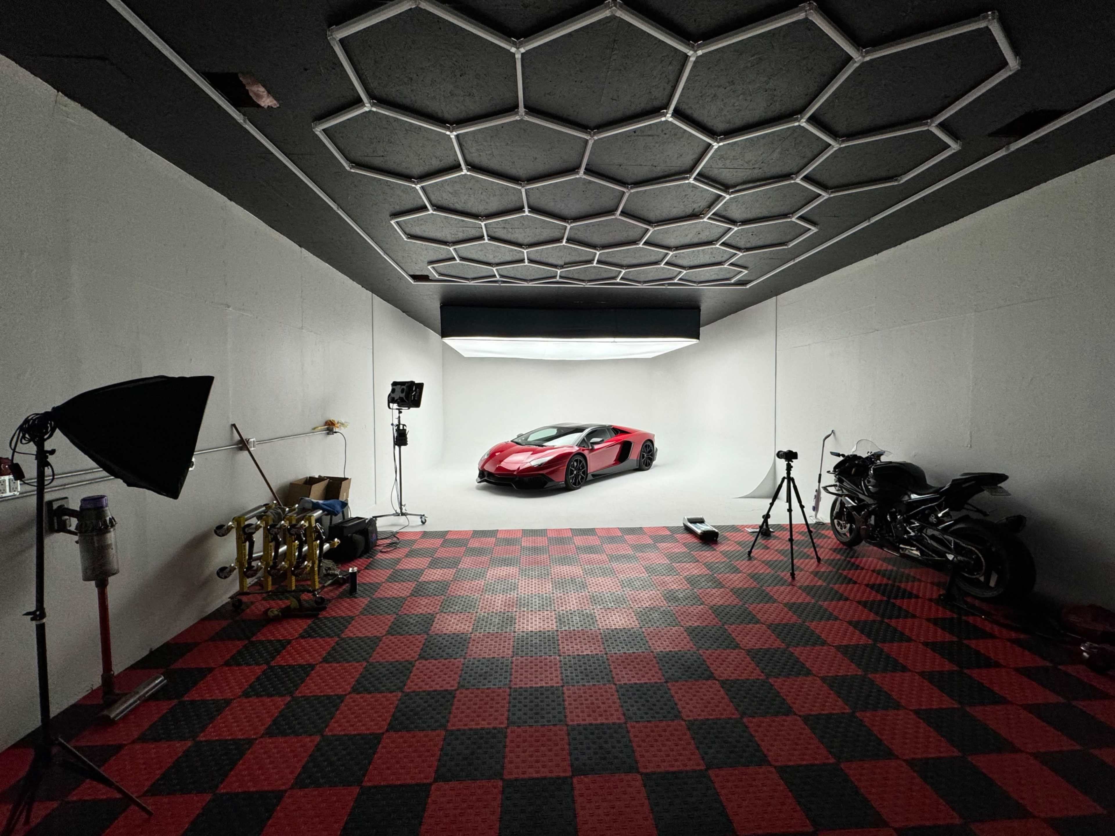 The image shows a spacious, well-lit garage featuring a red and black sports car, two motorcycles, and studio lighting equipment against a checkered floor.