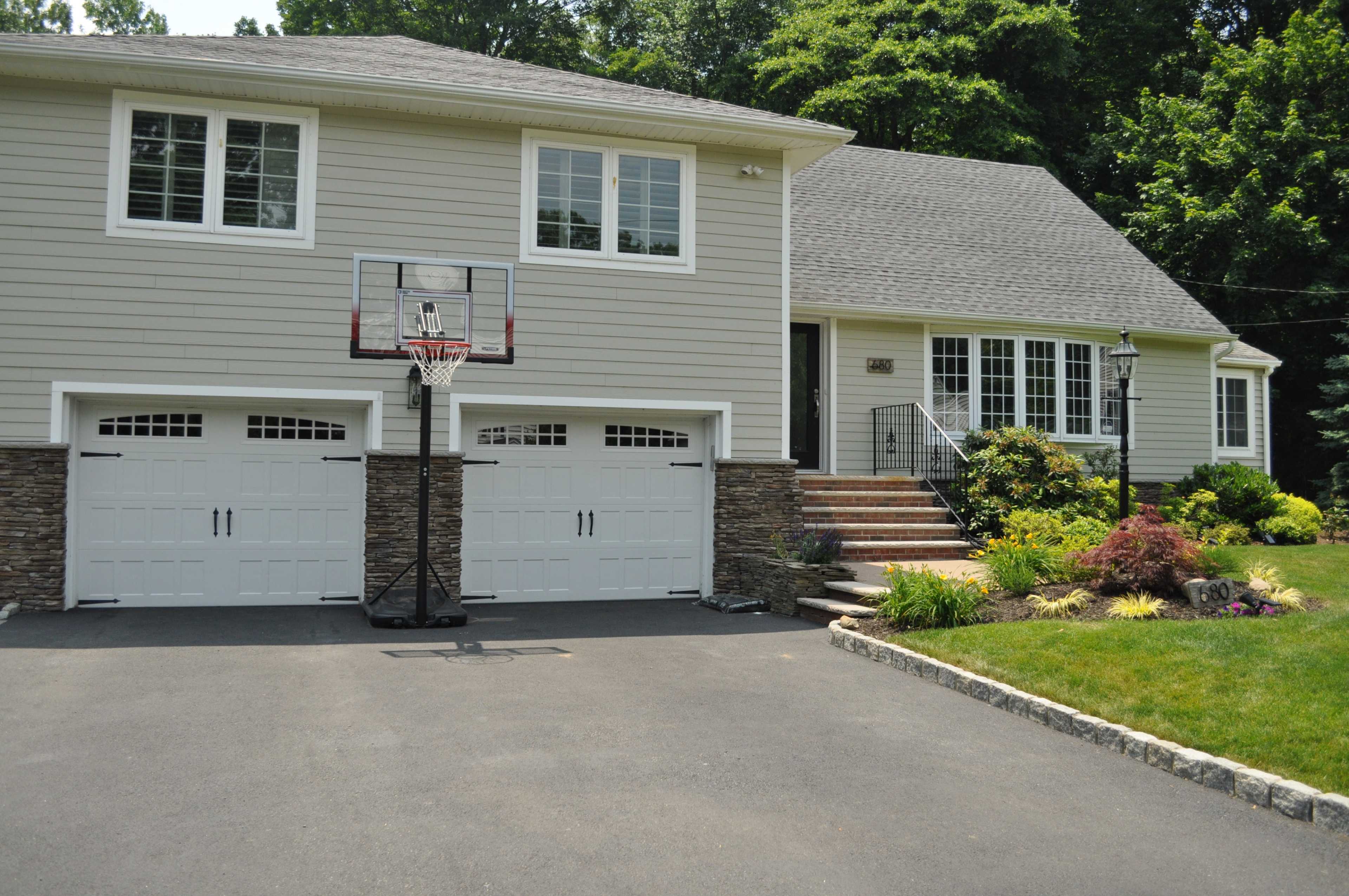 A house with a gray exterior, featuring a basketball hoop in the driveway and two garage doors surrounded by landscaped greenery.