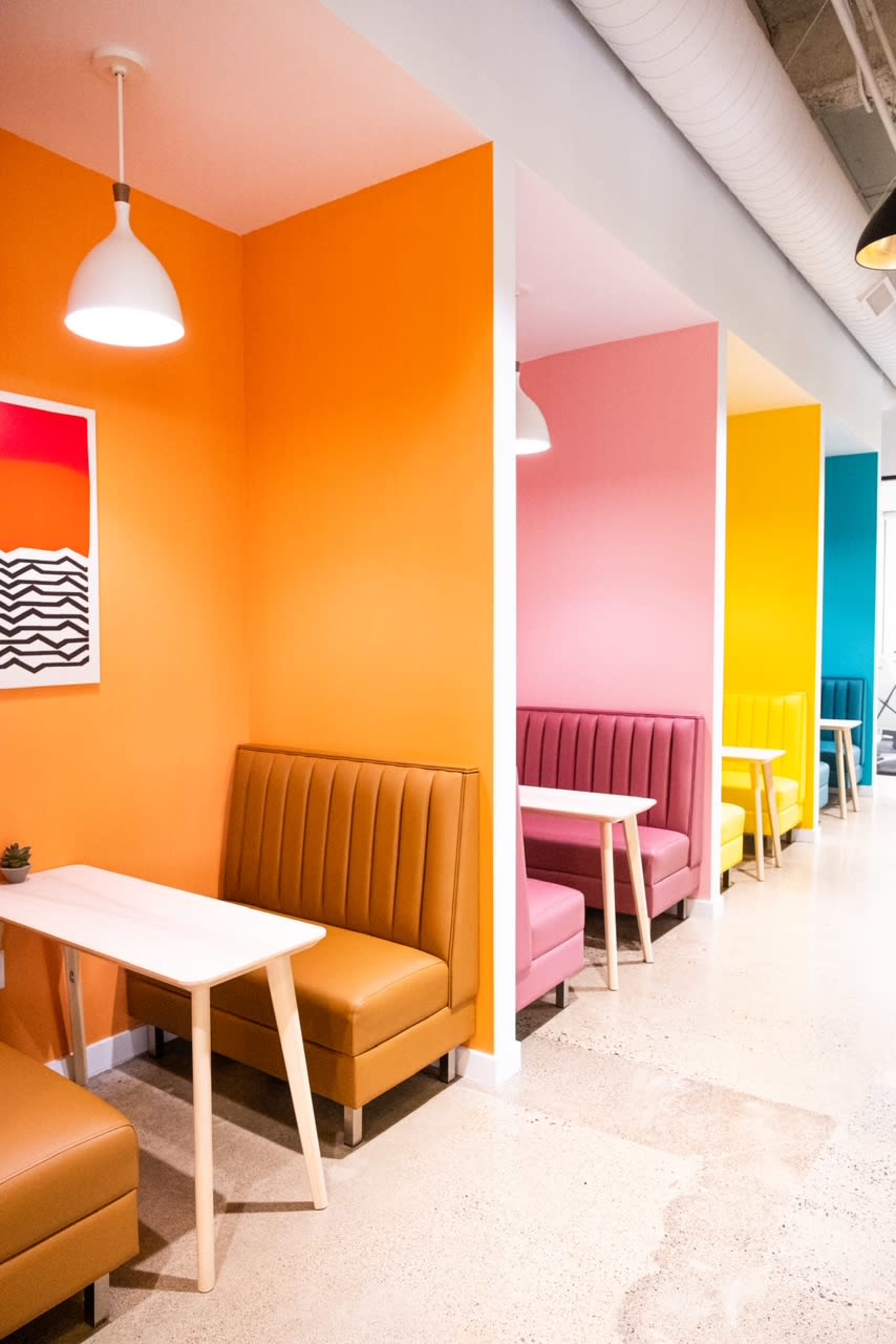 The image shows a brightly colored café interior with booths in orange, pink, and yellow walls, each featuring a small white table and brown leather seating.