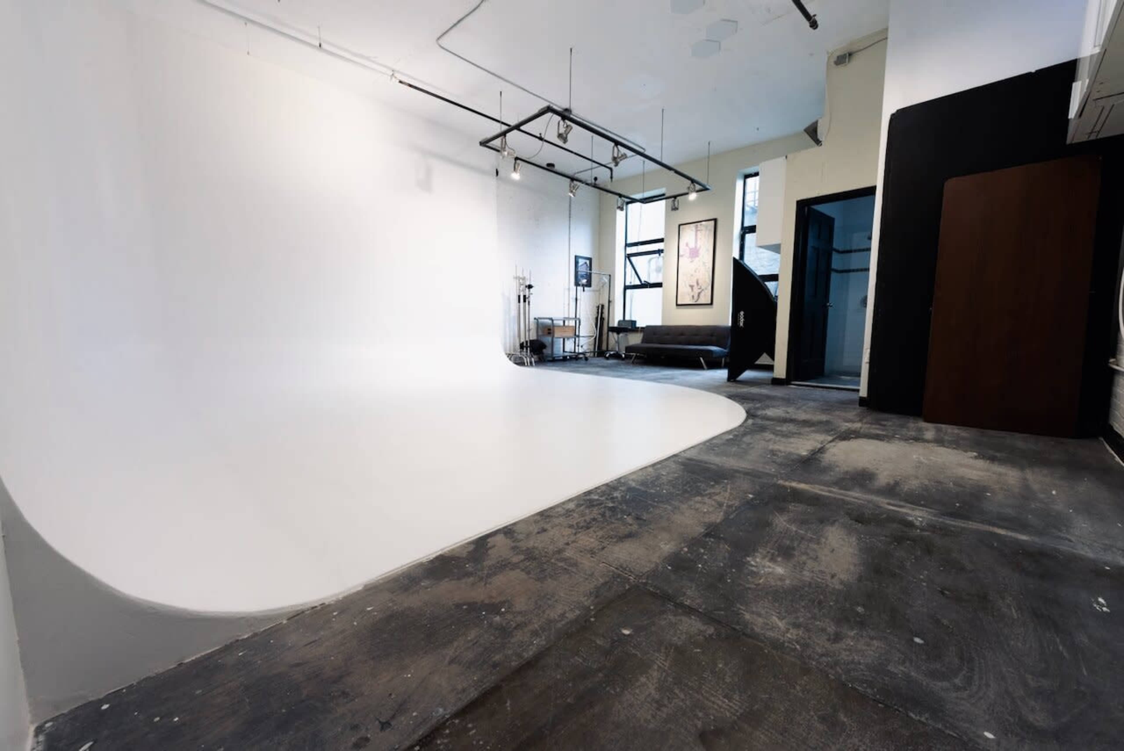 A spacious photography studio with a seamless white backdrop, exposed concrete floors, and minimal furnishings.