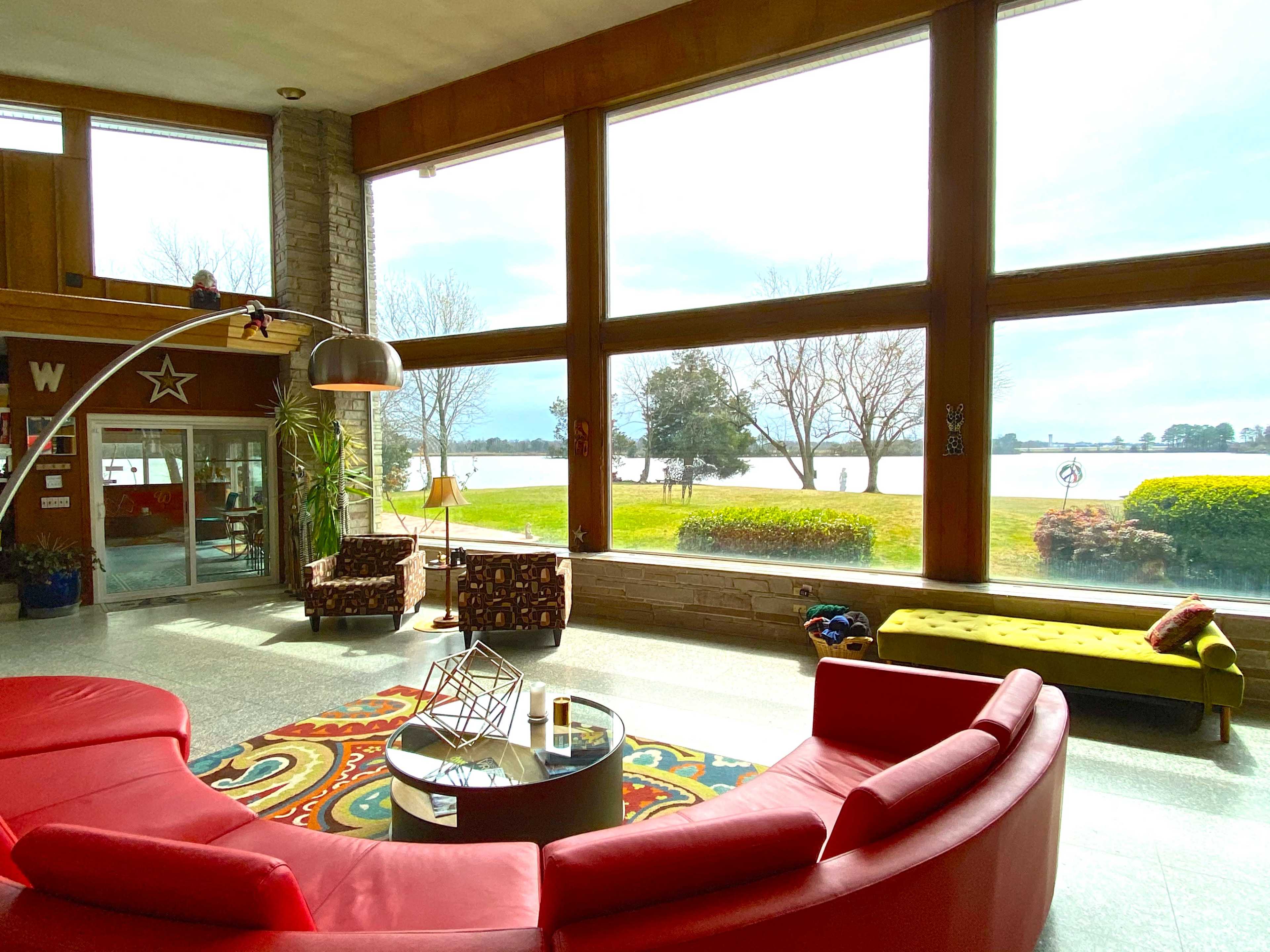 A spacious living area features a red circular sofa, large windows with views of a lake, and colorful decor.