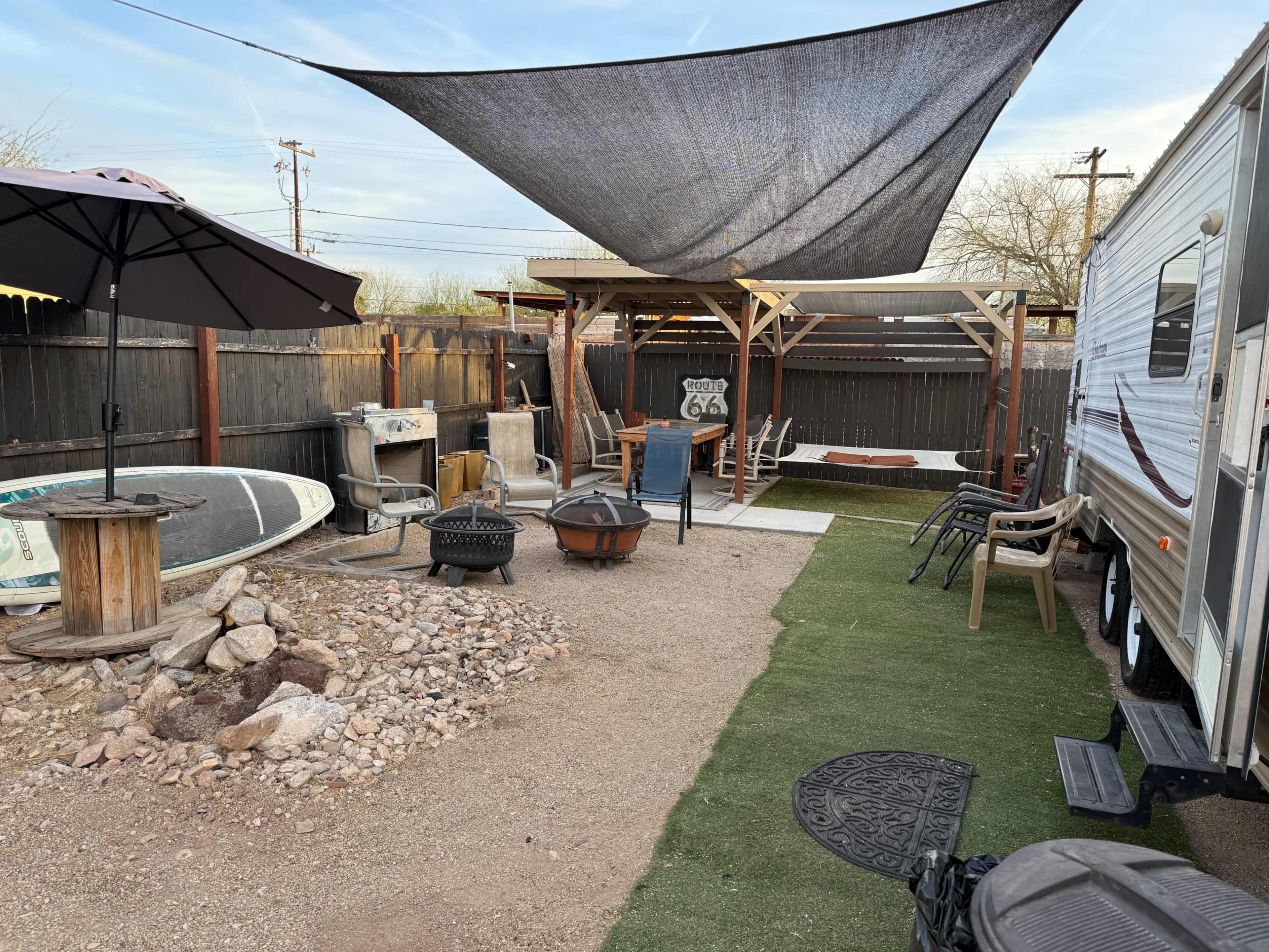 The image depicts a gravel yard with a shaded seating area, a travel trailer, and some outdoor furniture, including a fire pit and a paddleboard.