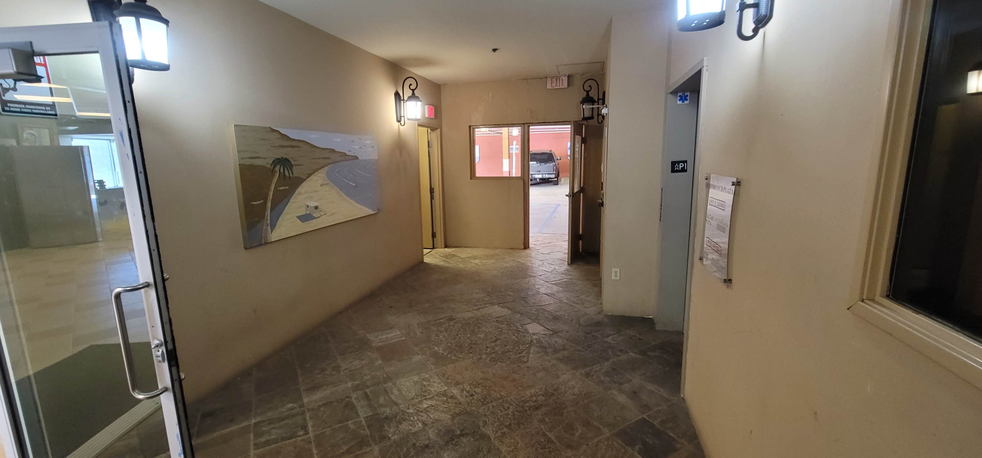 A hallway with tiled flooring, a painting on the wall, and doors leading to an outdoor area.