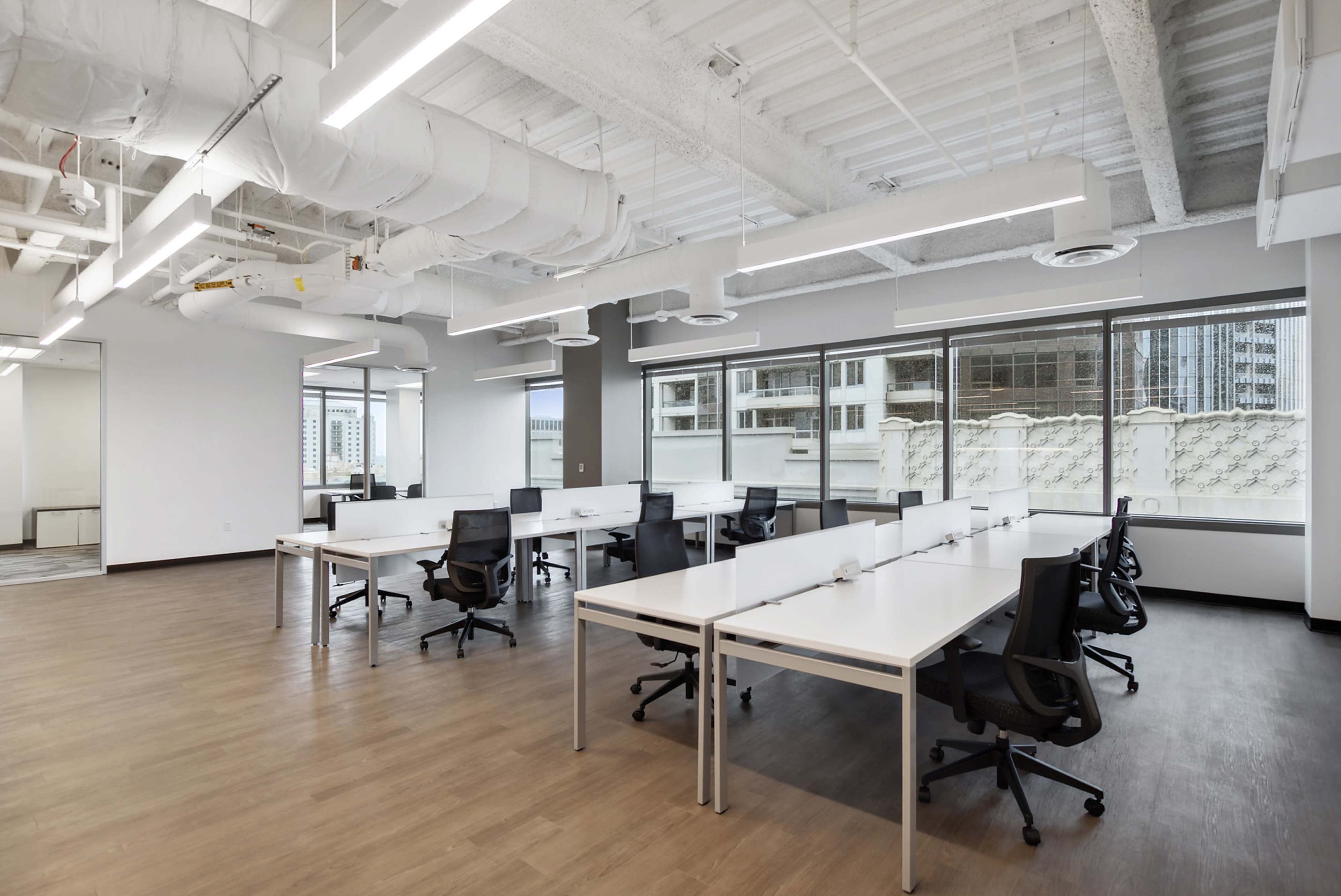 The image shows a modern office space featuring several white desks and black chairs arranged in an open layout with large windows.