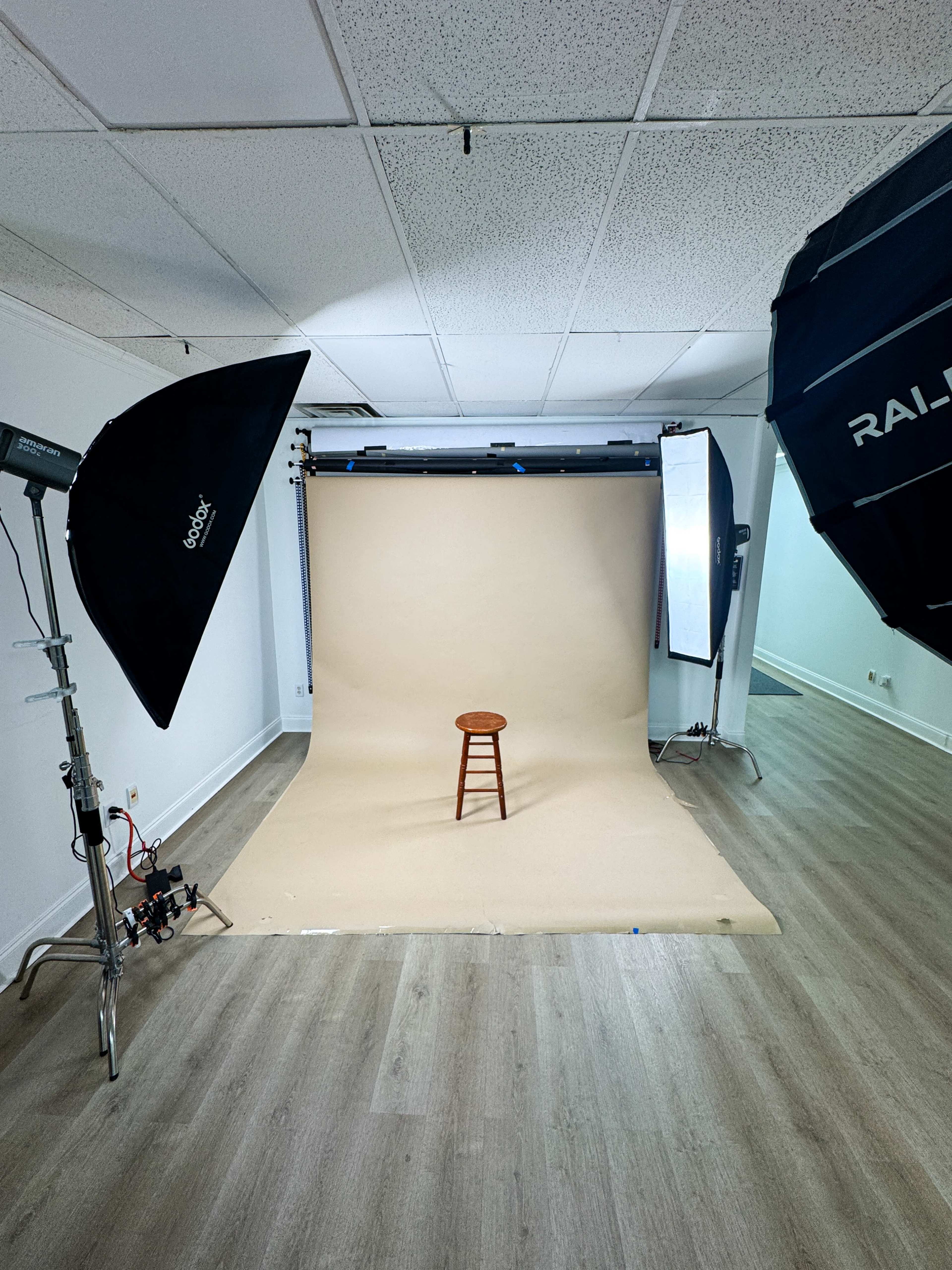 The image shows a photography studio setup featuring a plain backdrop, a wooden stool in the center, and lighting equipment positioned around the scene.
