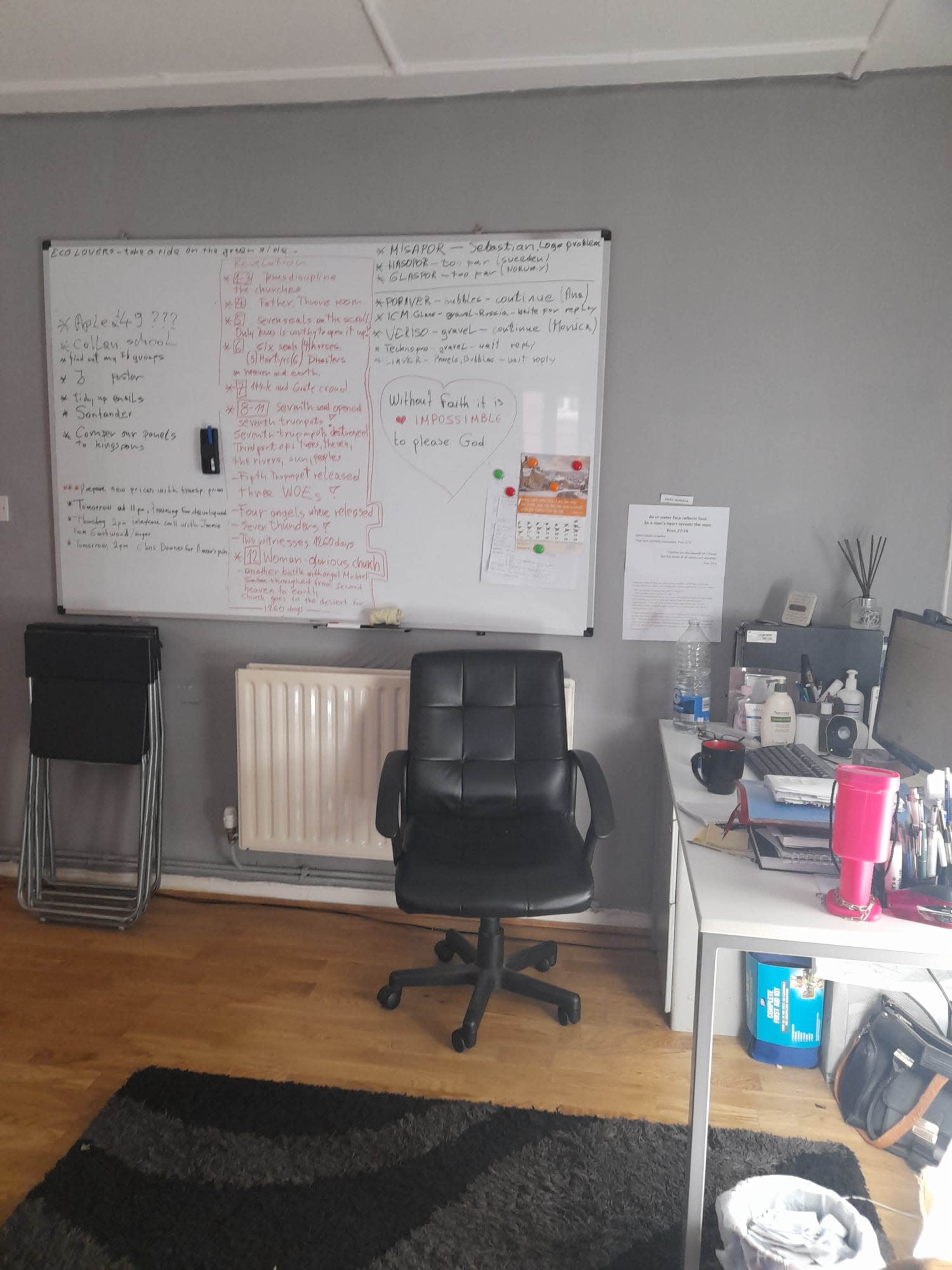 A home office with a black rolling chair, a whiteboard covered in notes, a desk with various items, and a radiator on the wall.