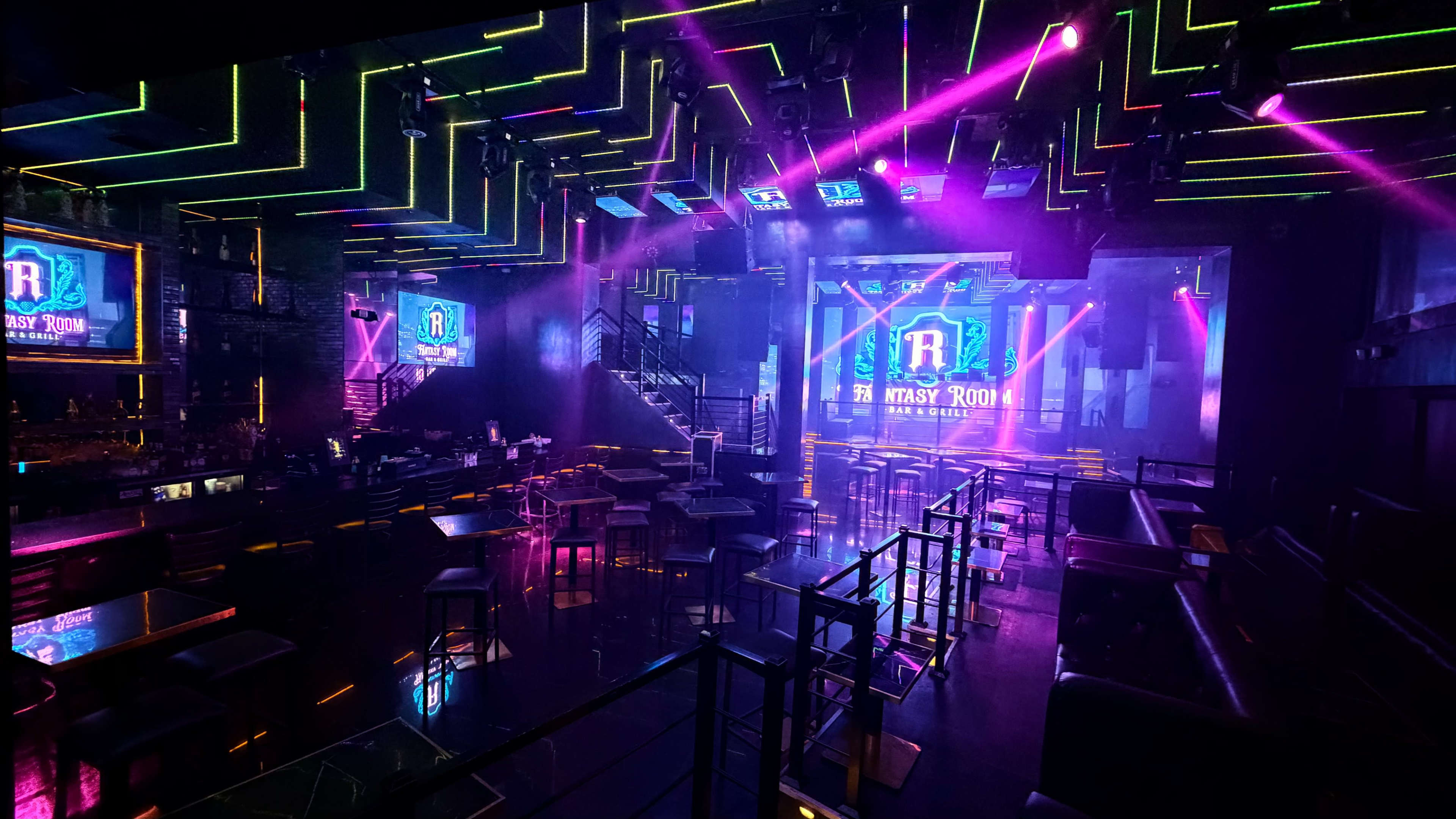 The image shows a vibrant nightclub interior with colorful neon lights illuminating tables and a stage.