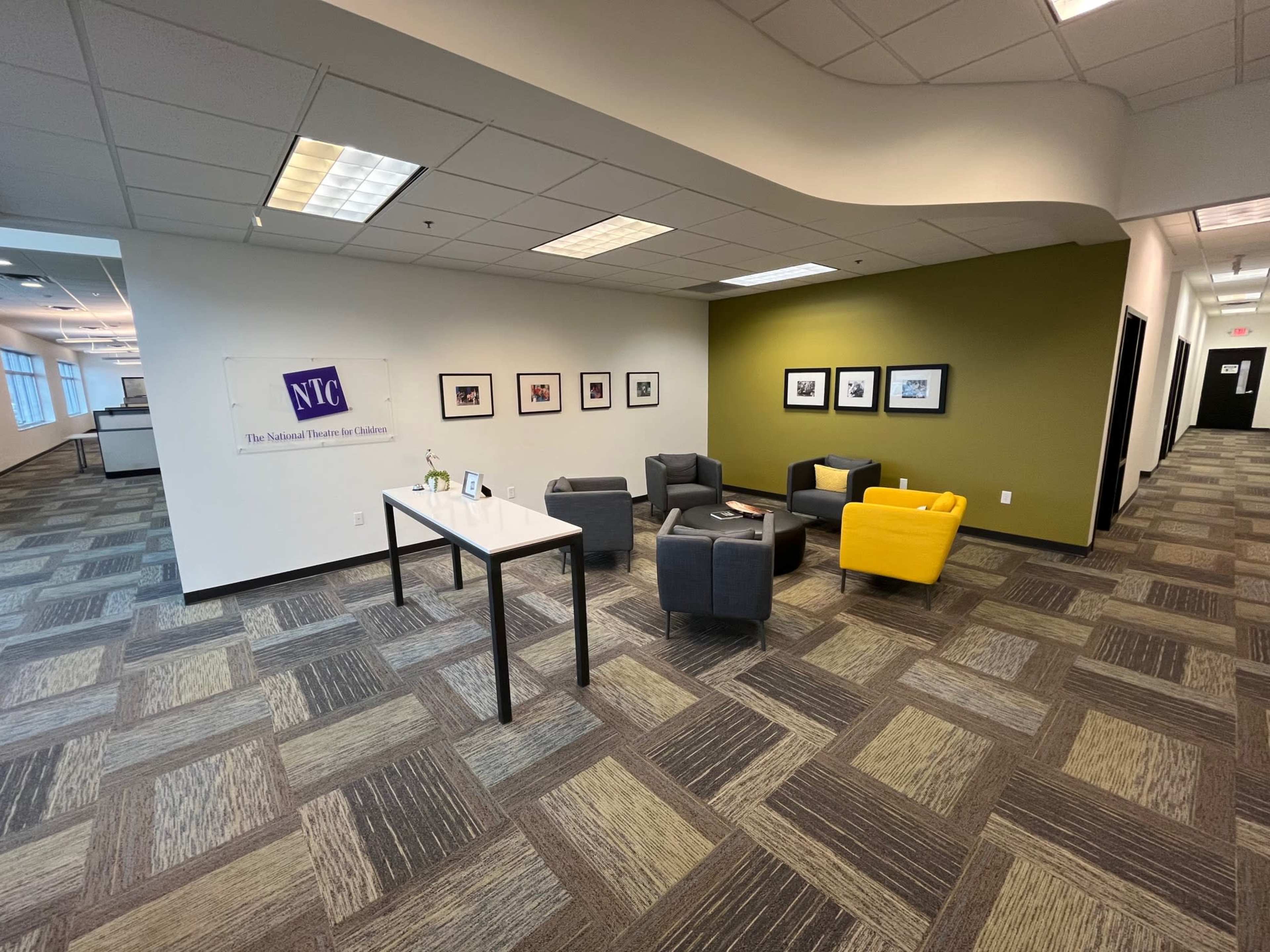 The image shows a modern office lobby with gray and yellow chairs arranged around a small table, framed pictures on the wall, and a logo banner on the left.