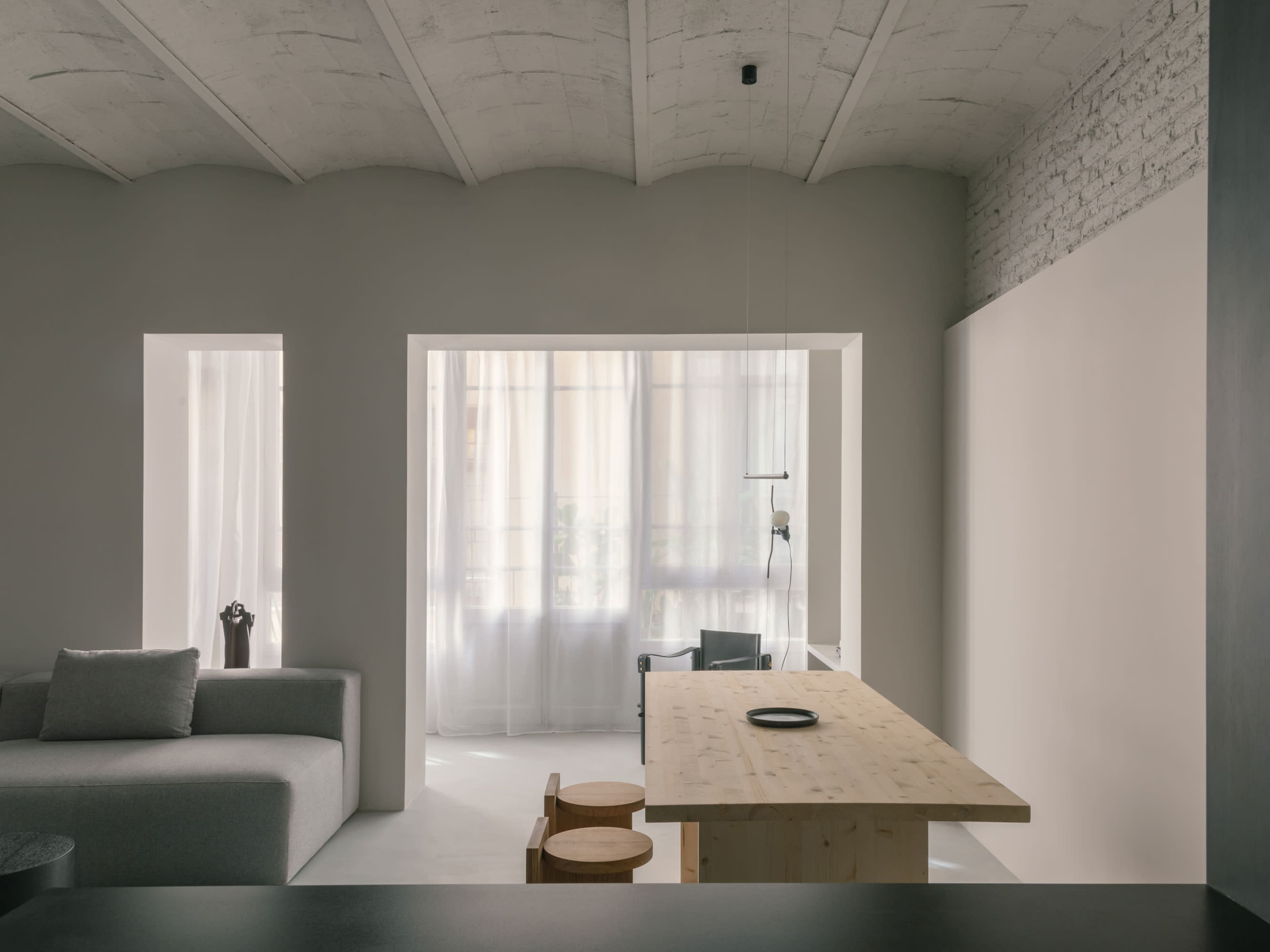 The image shows a modern, minimalist interior featuring a large wooden table, a gray sofa, and bright curtains allowing natural light to filter into the room.