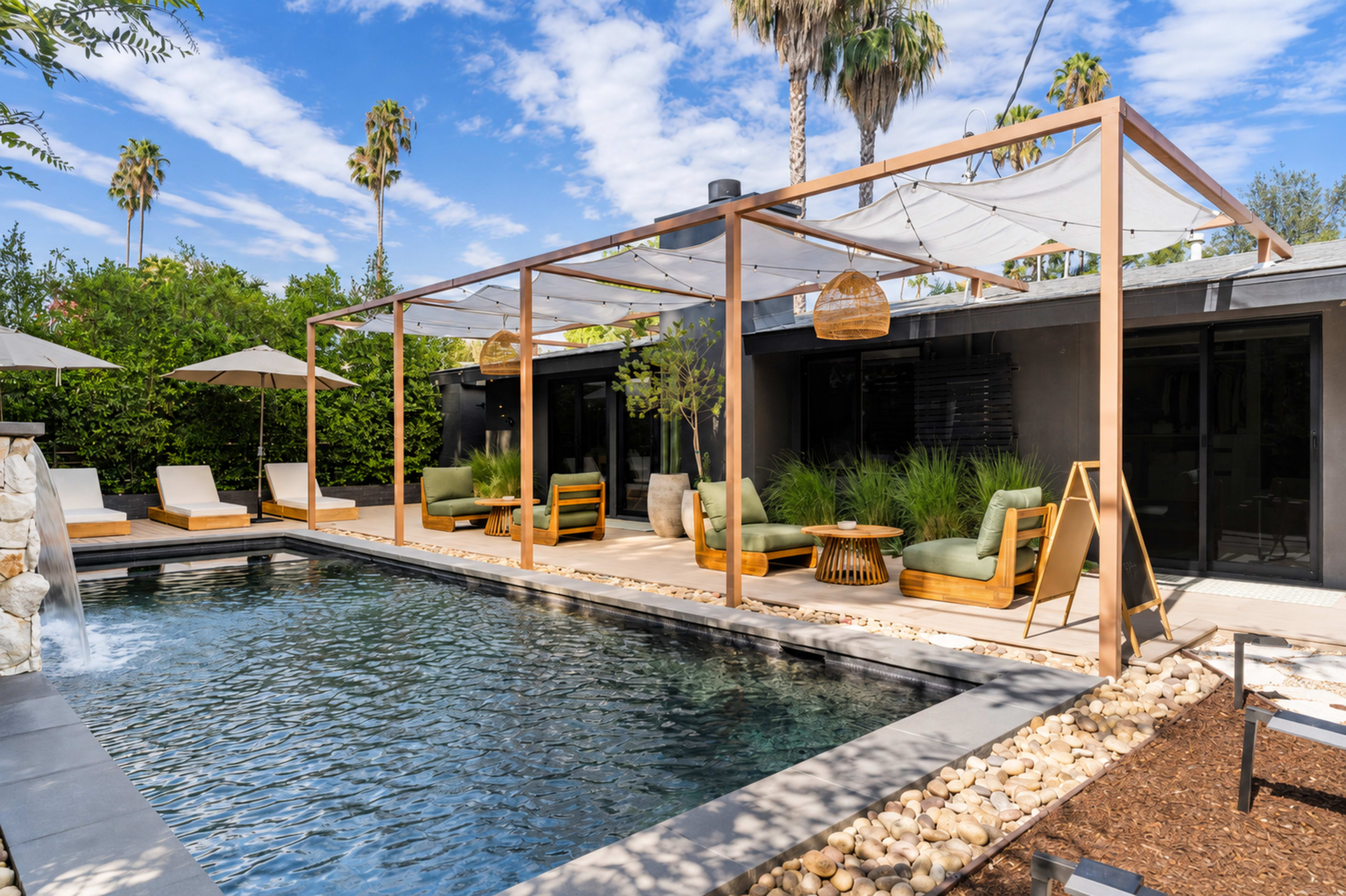 The image shows a modern outdoor pool area with lounge chairs, a pergola, and palm trees under a blue sky.