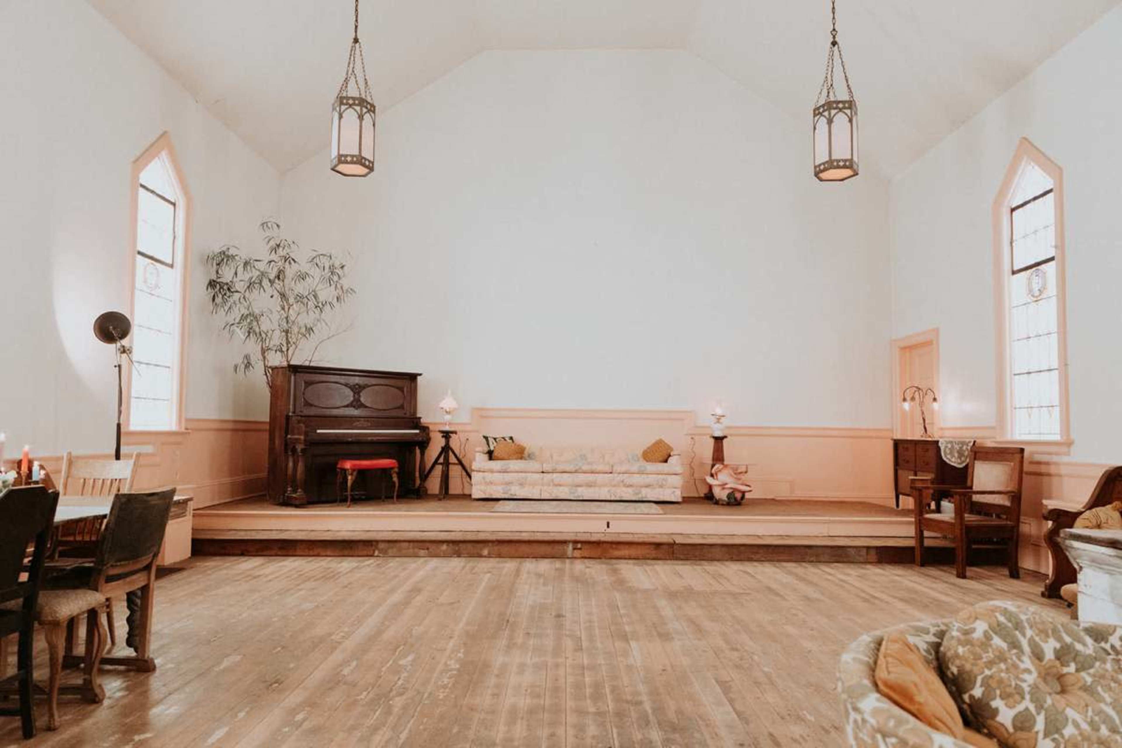 The image shows an airy interior of a spacious room with wooden floors, a vintage sofa, and an upright piano, framed by large windows and warm lighting.