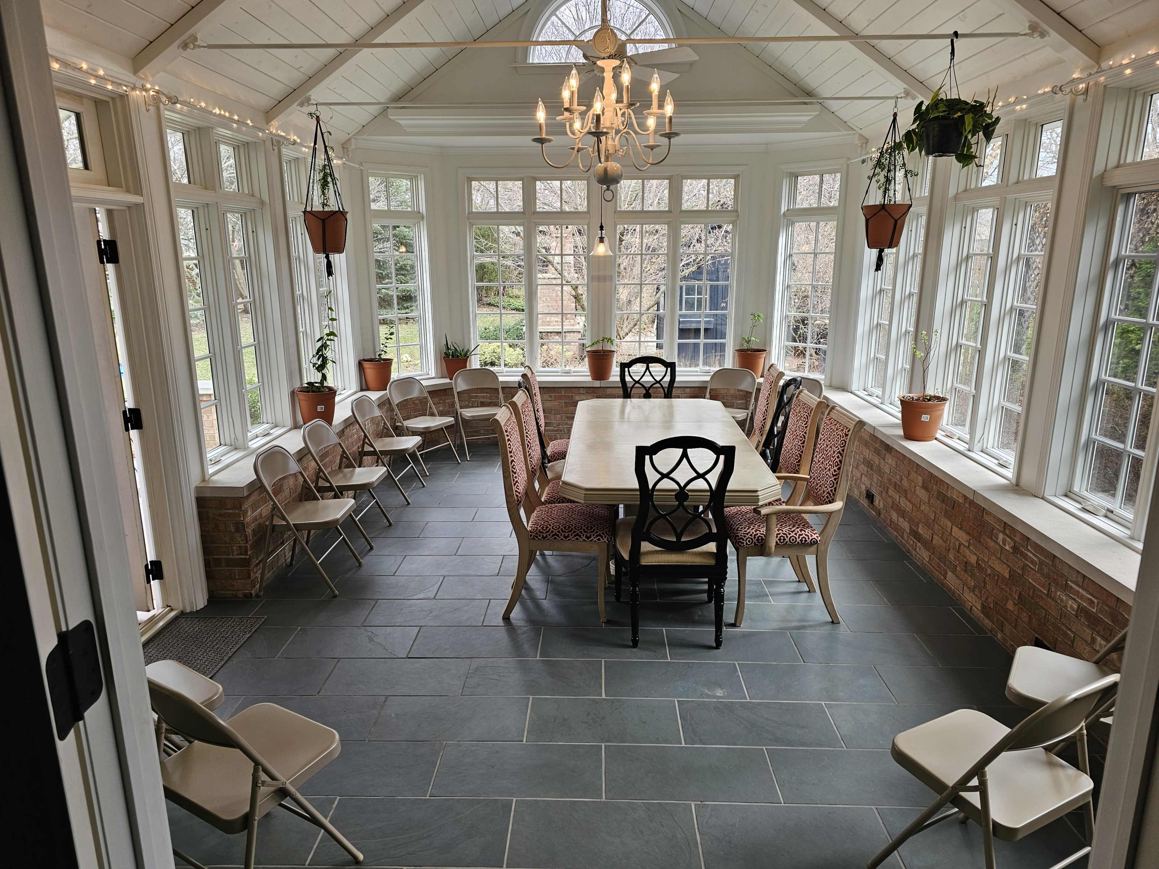 A sunroom with a large table surrounded by chairs, featuring large windows that provide views of the outdoor garden.