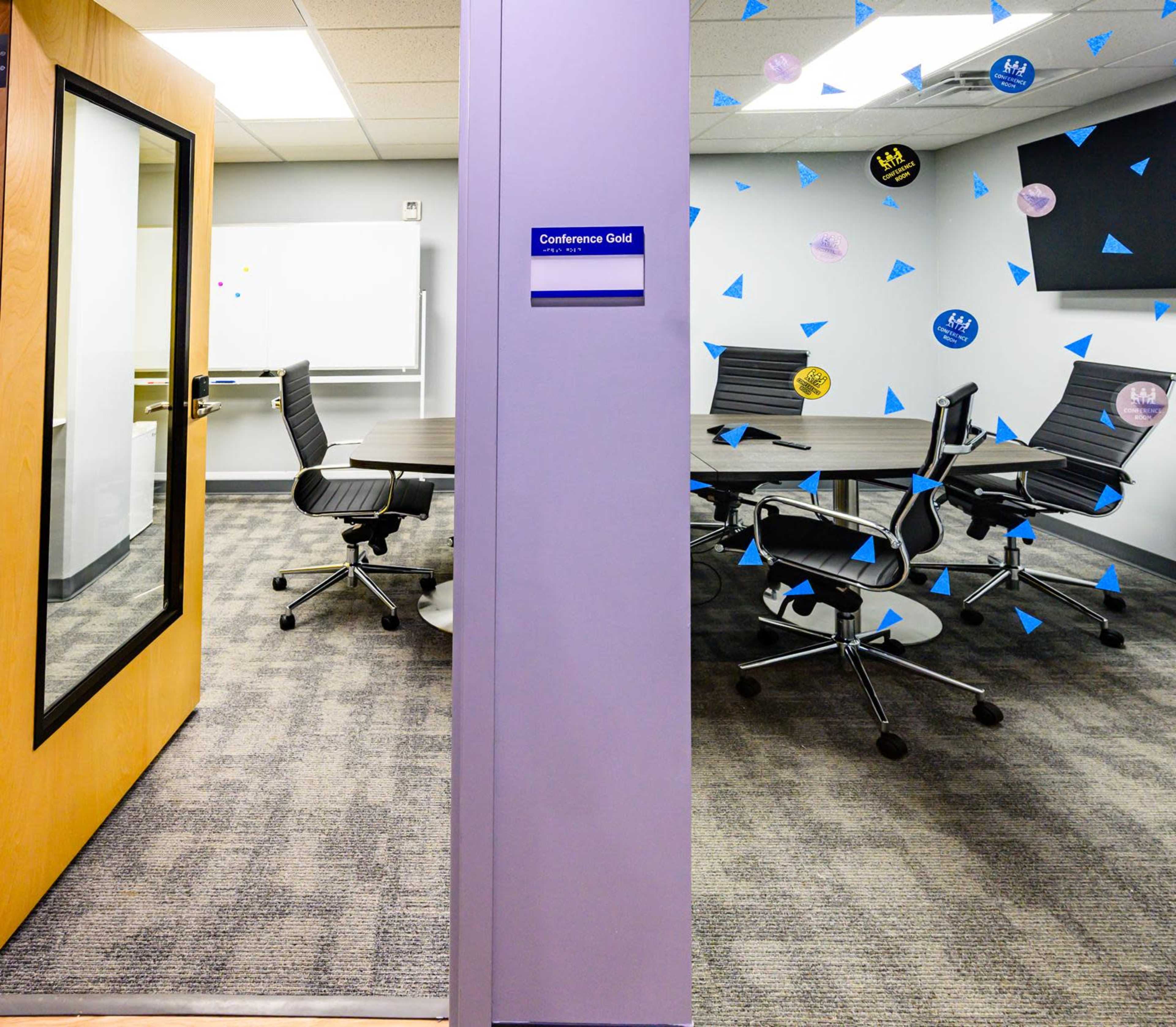 The image shows the entrance to a conference room labeled "Conference Gold," featuring a modern meeting space with chairs and a table visible inside.