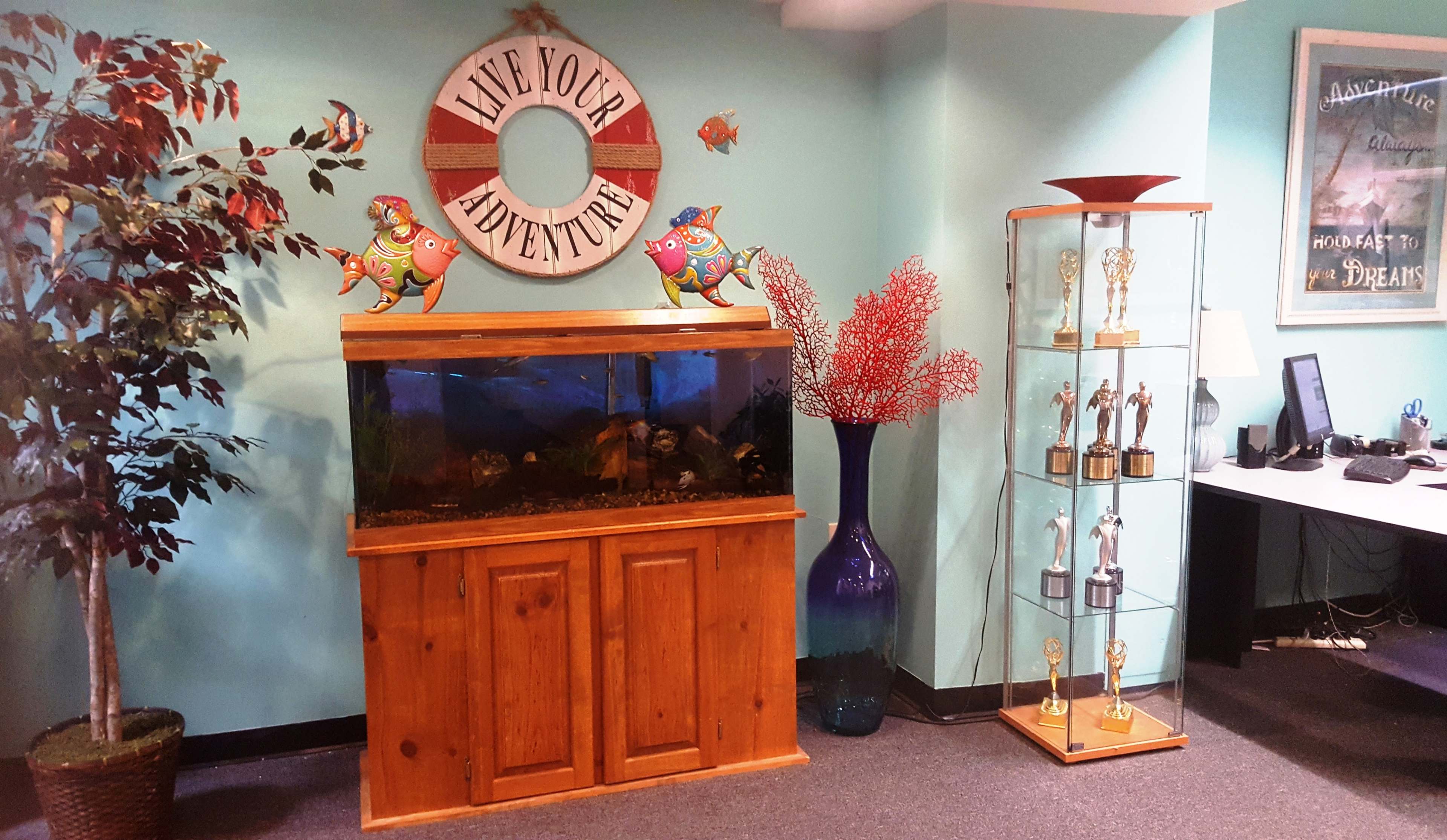 The image shows a wooden aquarium filled with fish, flanked by decorative plants, a trophy display, and a wall featuring a nautical-themed sign that reads "Live Your Adventure."