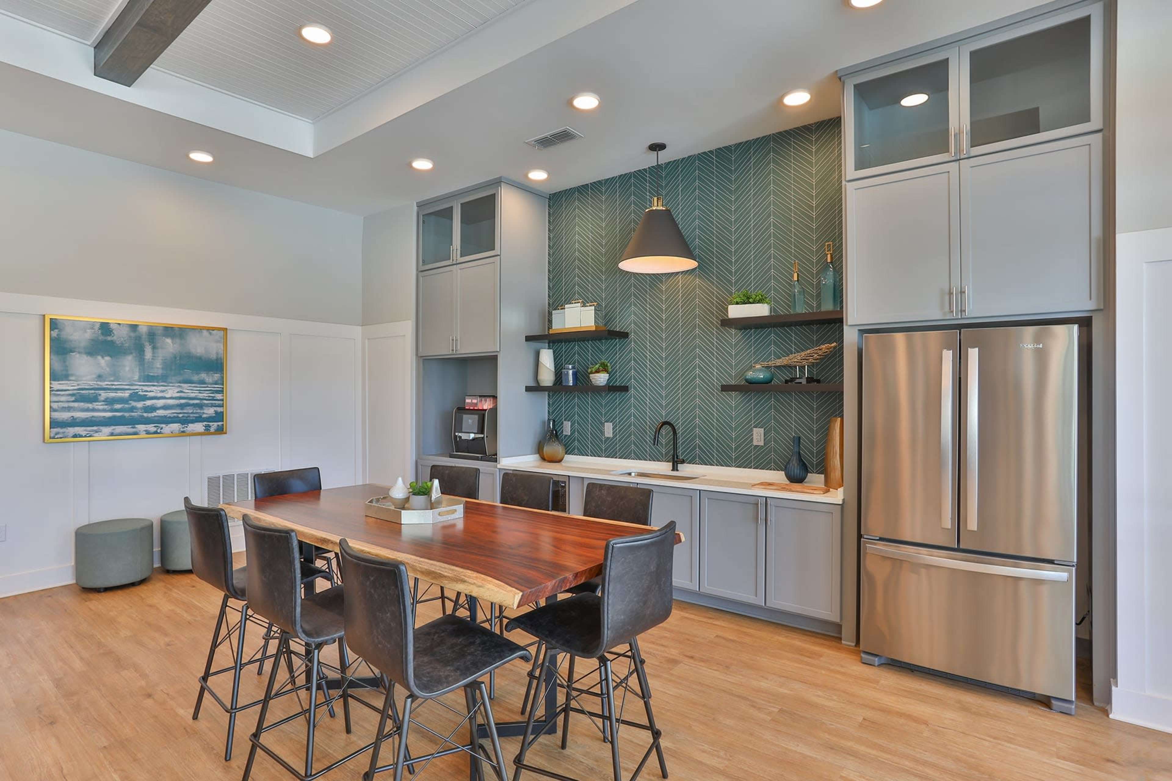 A modern kitchen with a wooden dining table, gray cabinets, and a stainless steel refrigerator, featuring a patterned green backsplash.