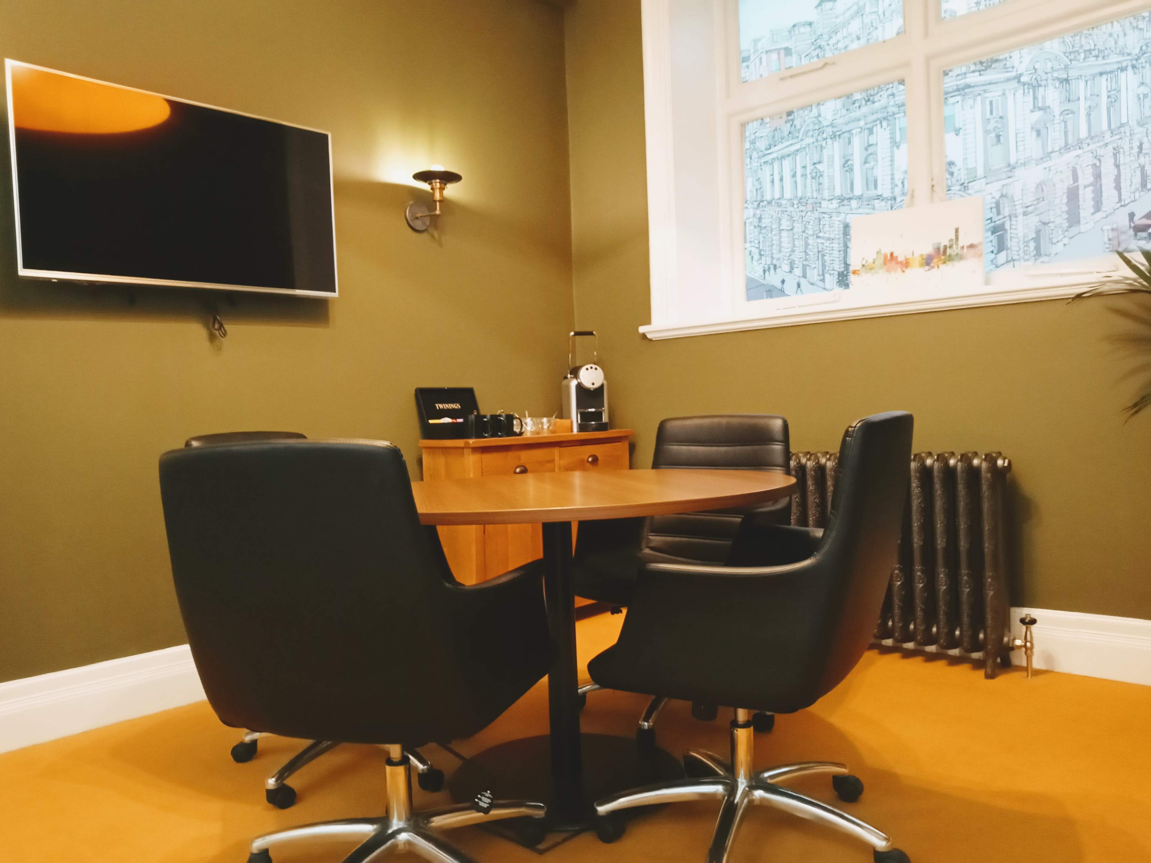 A small conference room features a round table with three black chairs, a television mounted on the wall, and a window with a view of a cityscape.