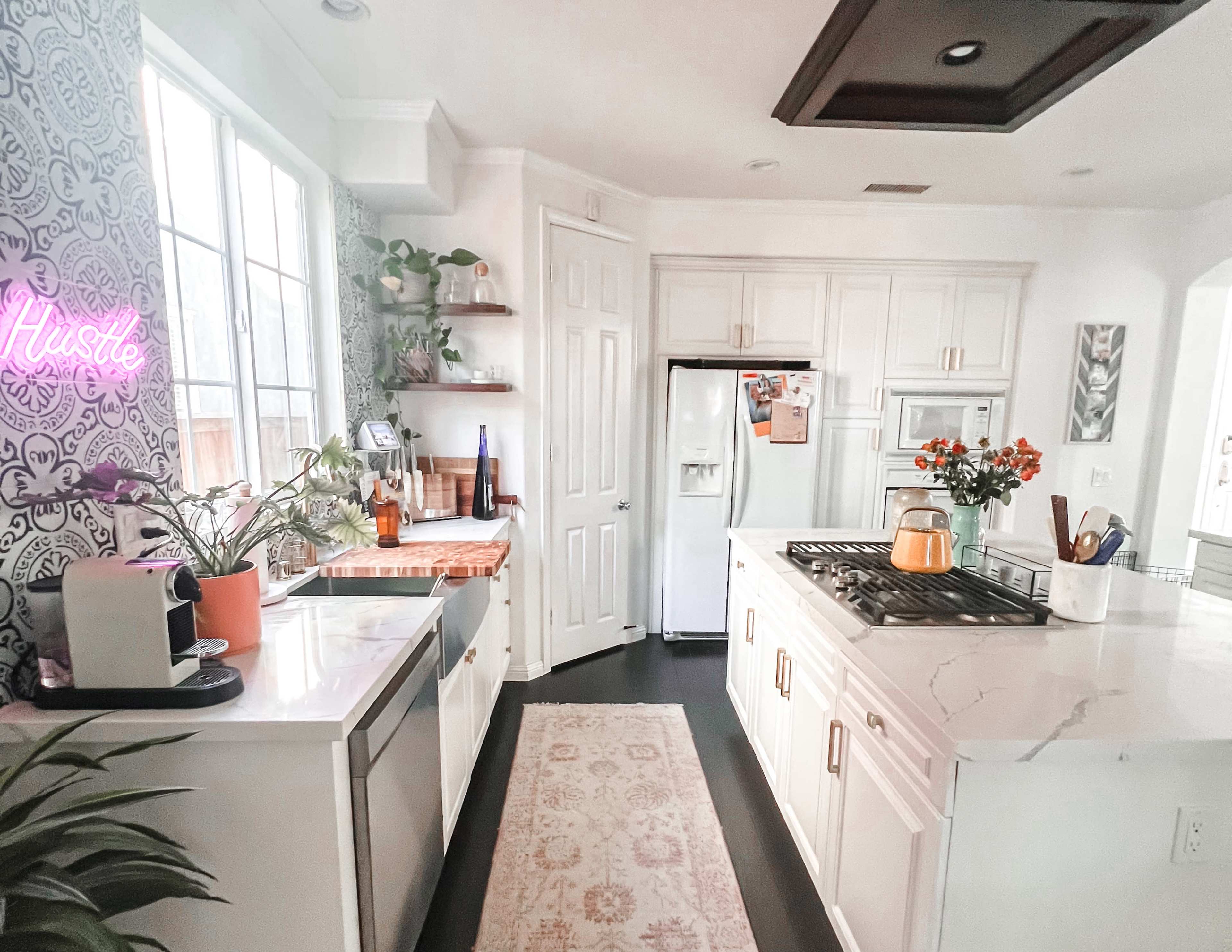 The image shows a modern kitchen with white cabinets, a gas stove, and a sleek countertop, featuring plants and a neon sign that says "Hustle."