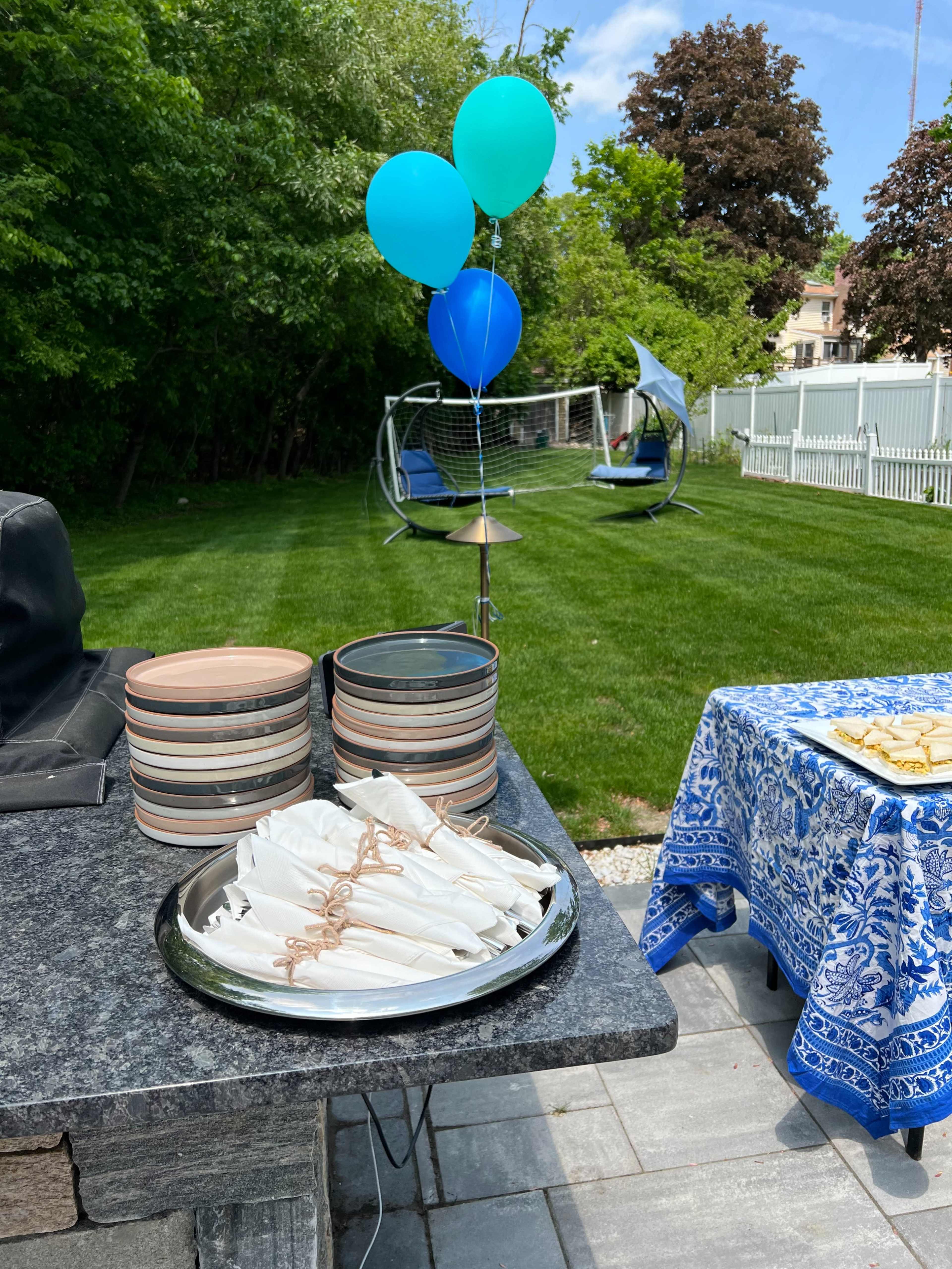 A neatly arranged outdoor setting features a table with plates, napkins, and balloons, alongside a grassy area with a swing and a hammock in the background.