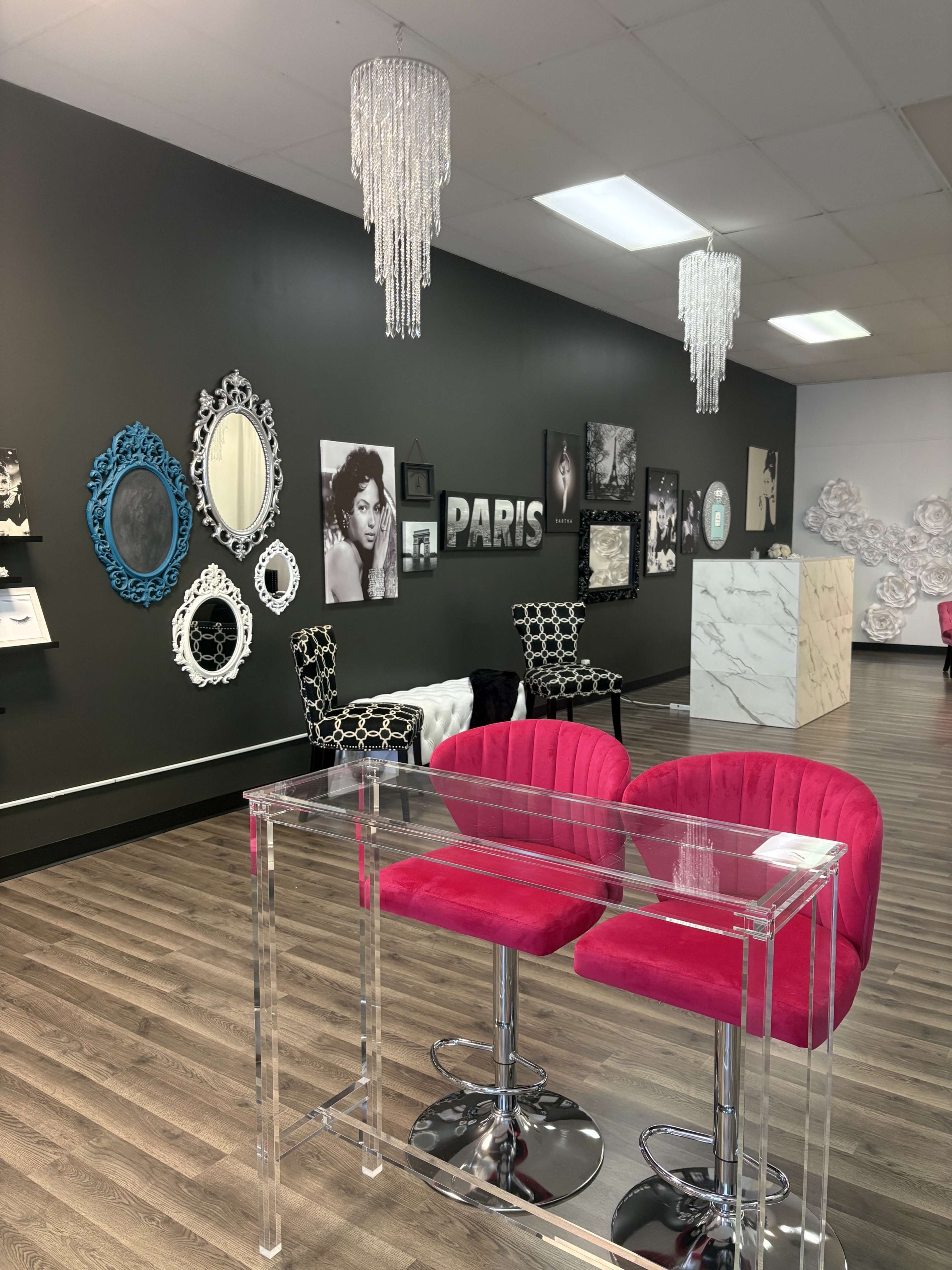 The image shows a stylish interior of a salon or boutique with pink bar stools, a clear acrylic table, framed black-and-white photos, and ornate mirrors displayed on the walls.