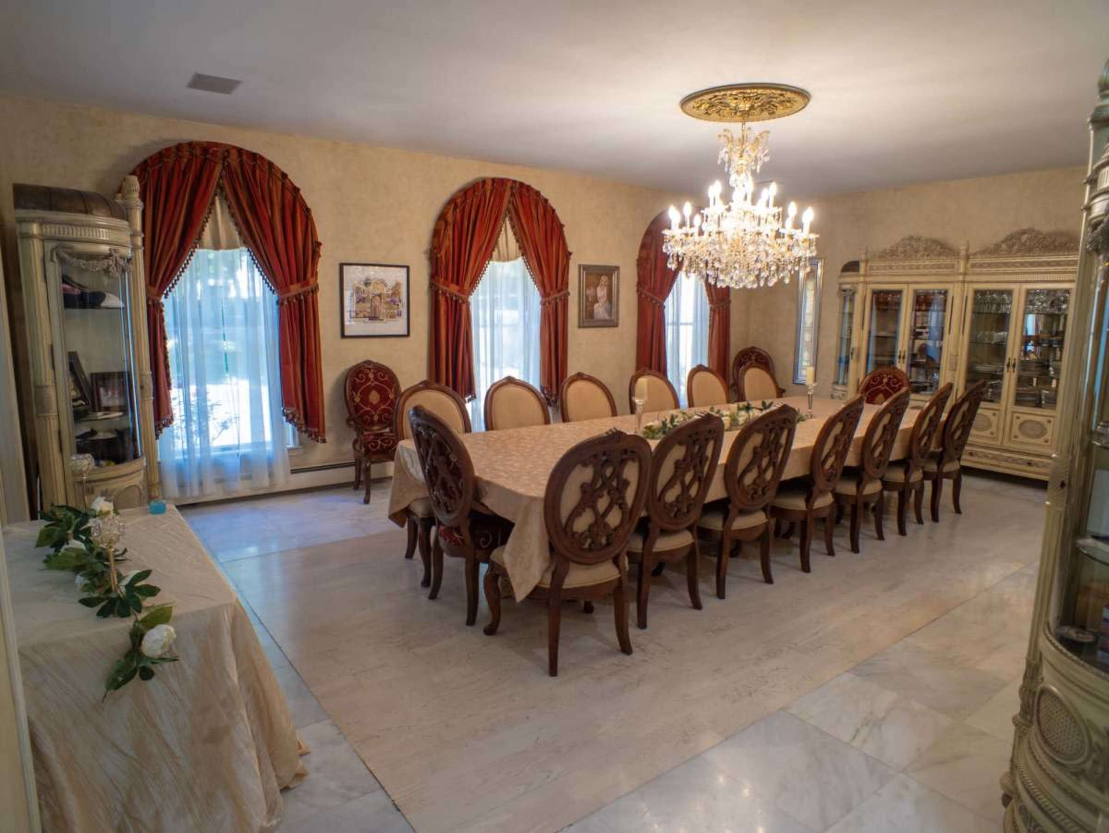 The dining room features a large table surrounded by ornate chairs, set beneath a chandelier and flanked by windows with draped red curtains.