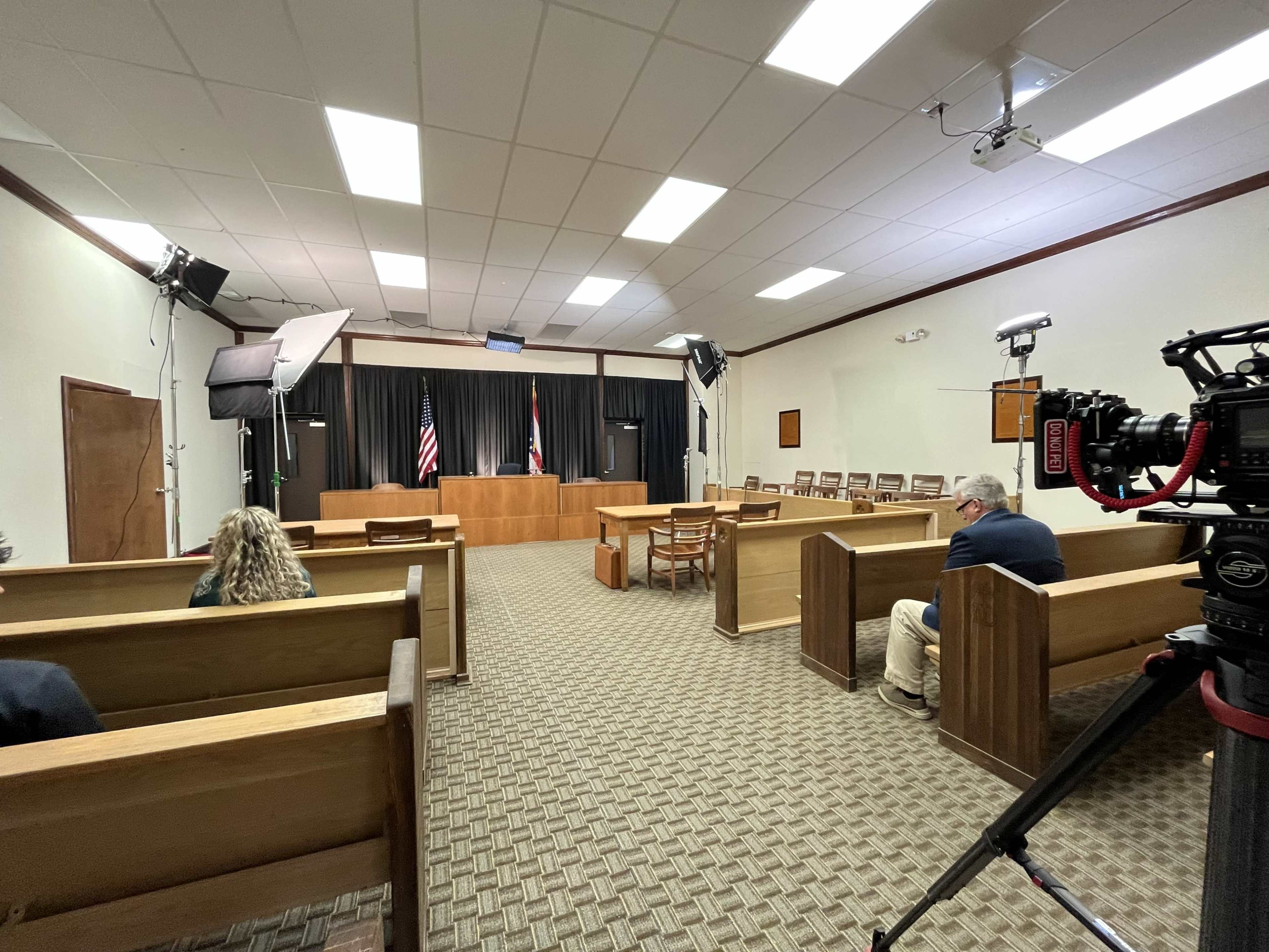 The image shows an empty courtroom with wooden benches, flags positioned at the front, and camera equipment set up for filming.
