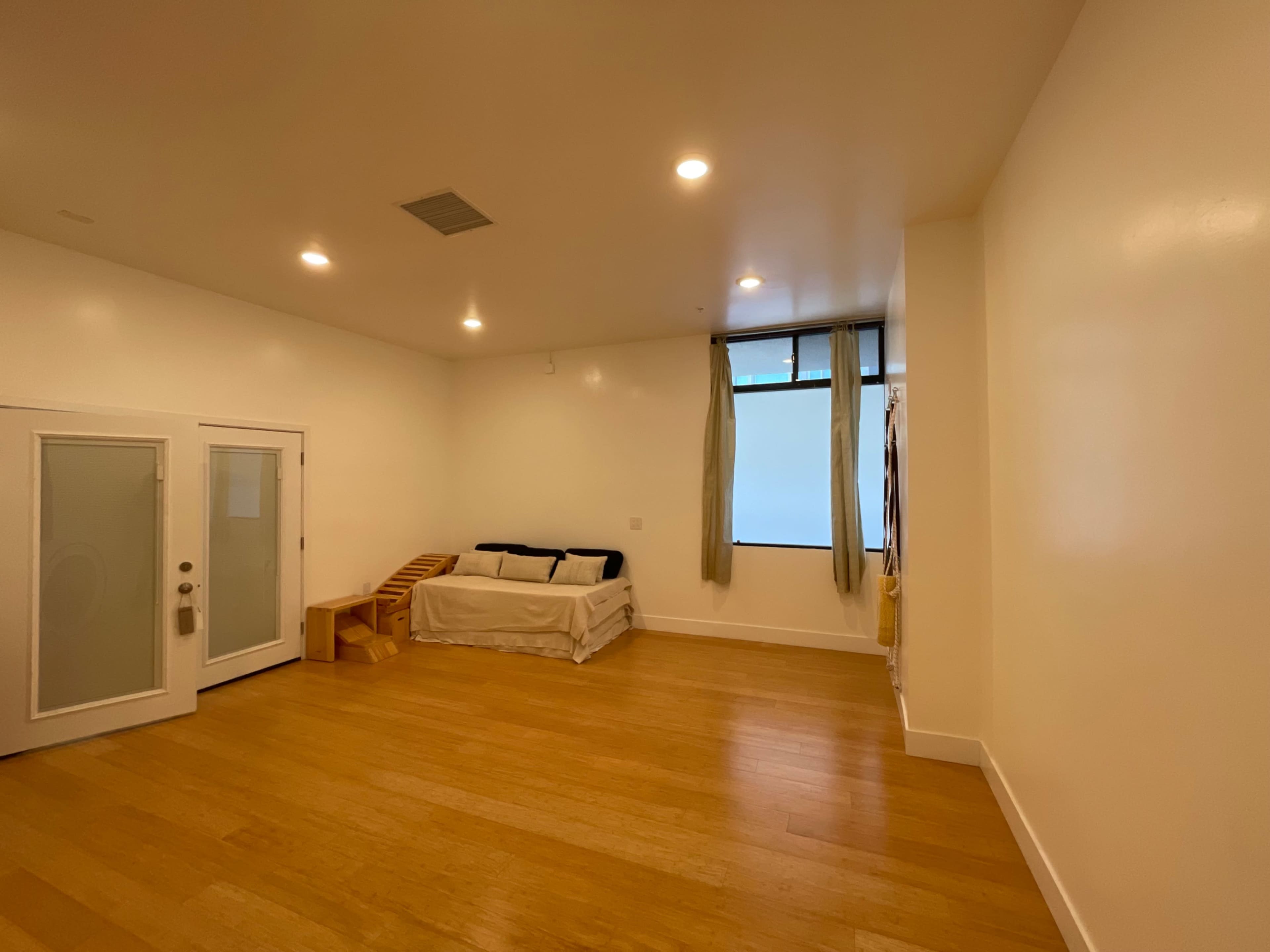 A spacious, well-lit room features a sofa, a wooden structure, and a large window with curtains.