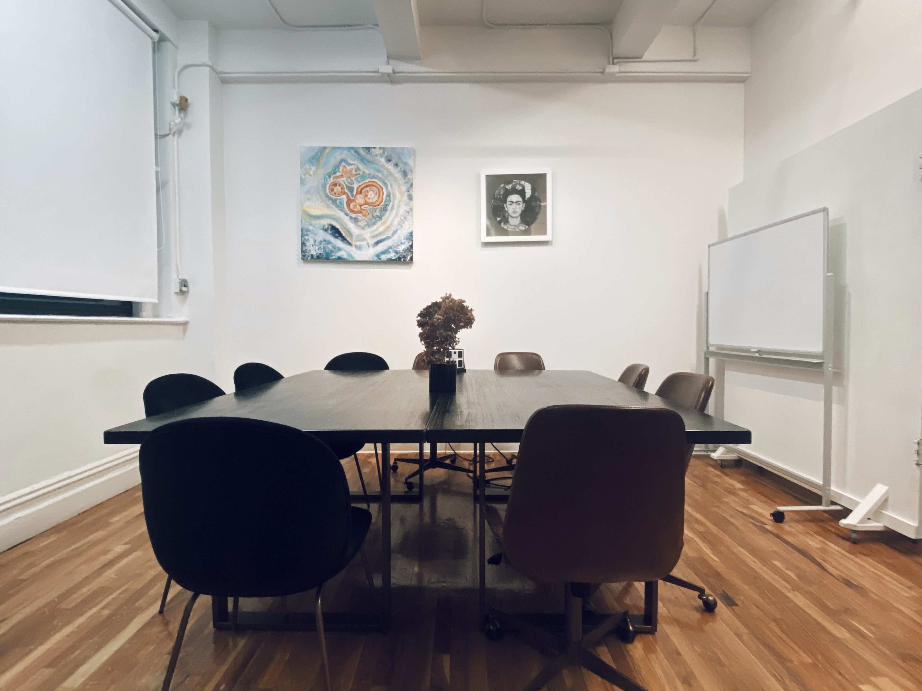 A conference room features a large wooden table surrounded by several chairs, with abstract art on the walls and a whiteboard on one side.