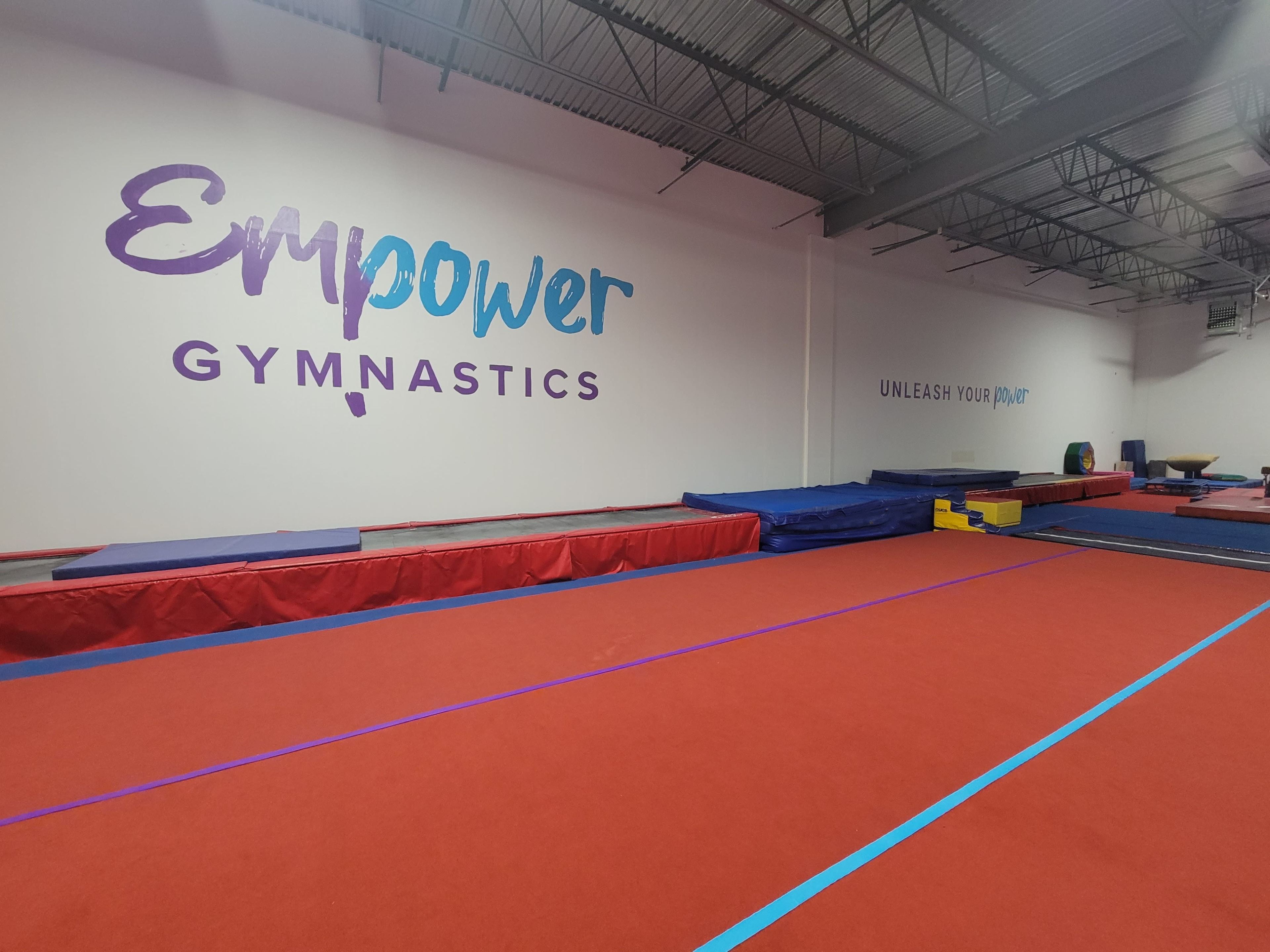 An indoor gymnastics facility with a red and blue mat layout and walls featuring the text "Empower Gymnastics" and "UNLEASH YOUR POWER."