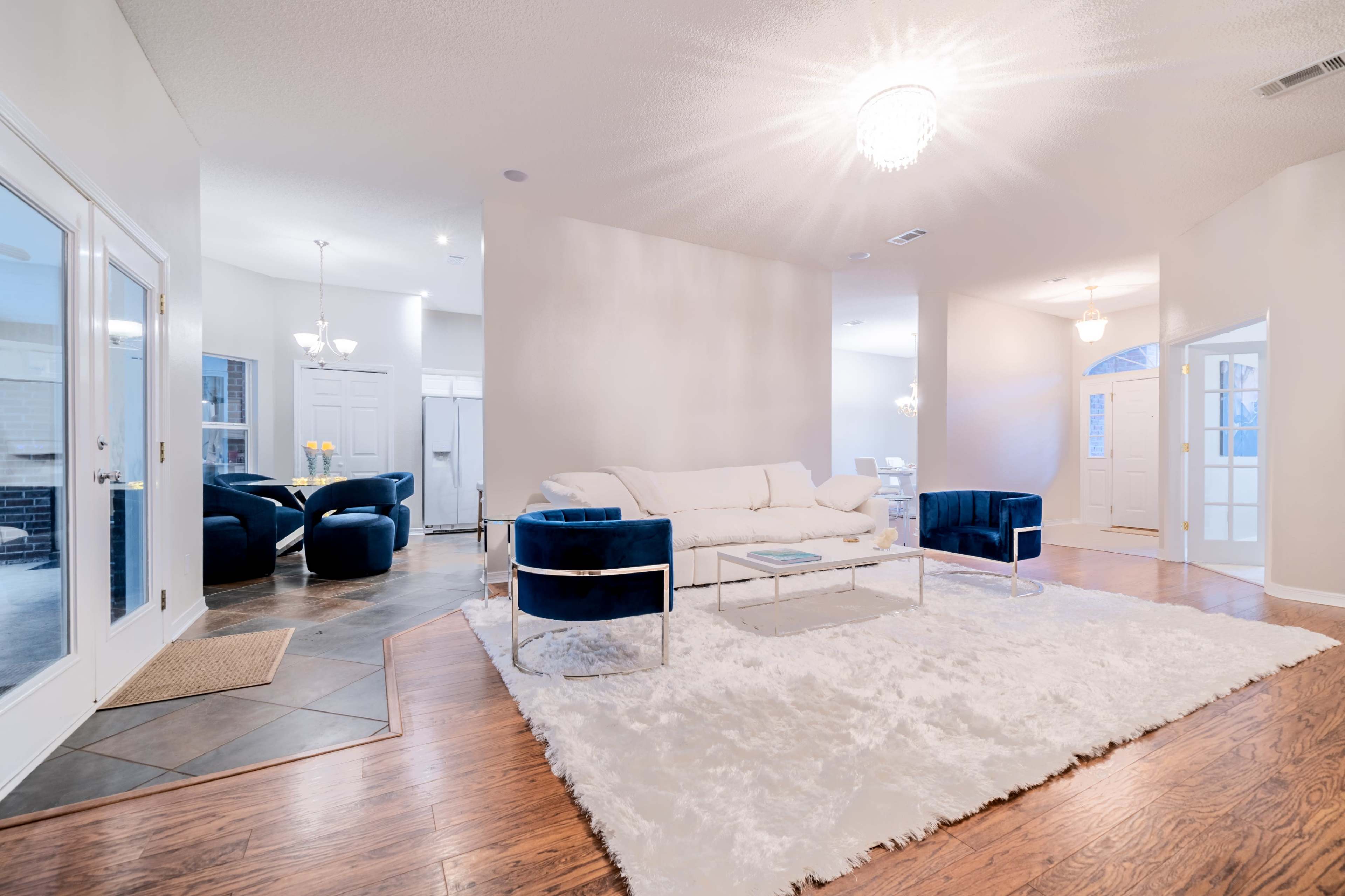 A modern living room with a white sofa, two blue velvet chairs, a glass coffee table, and a large white rug on hardwood floors.