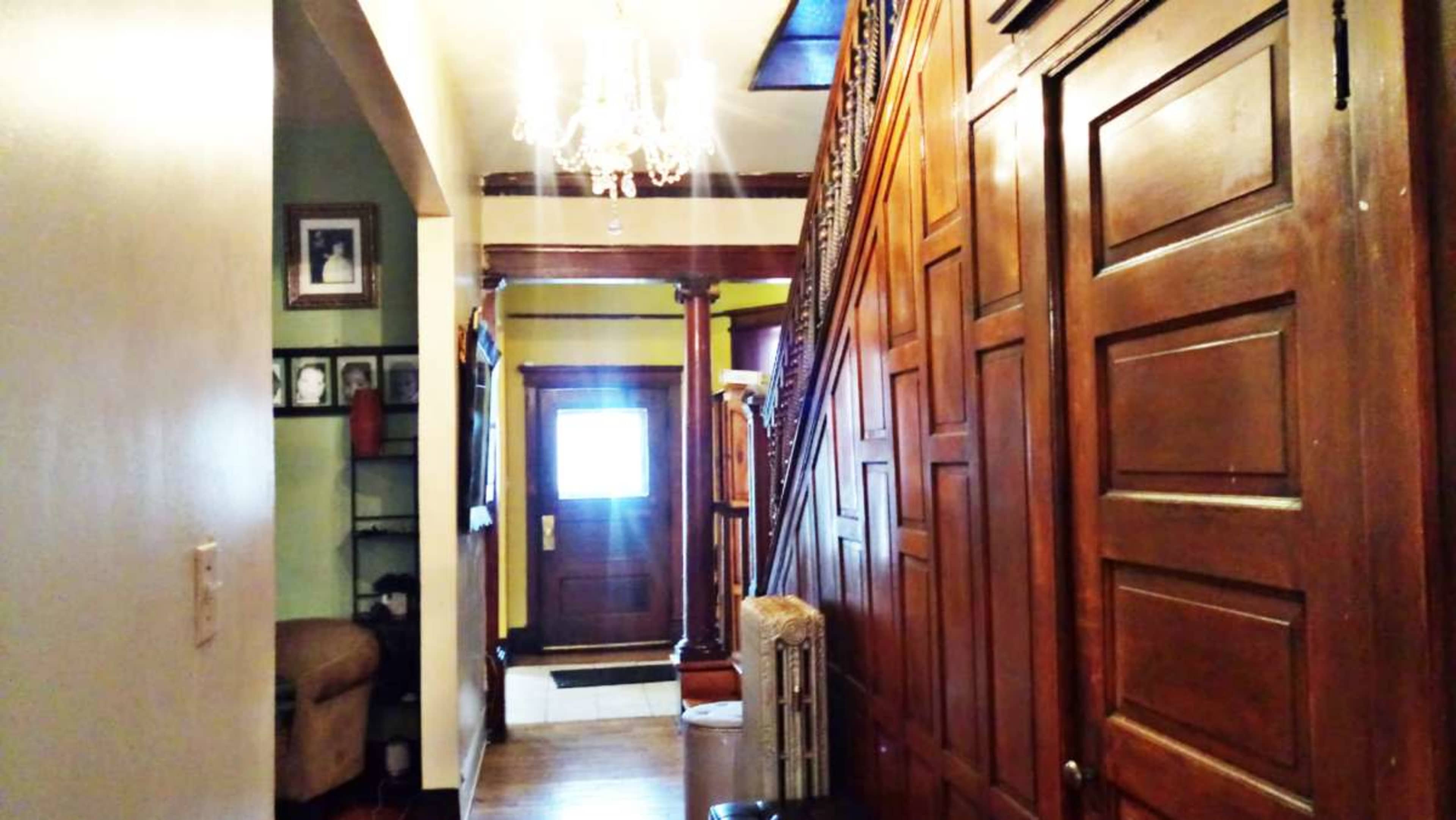 The image shows a hallway with wooden paneling, a staircase, and a door leading to the outside, illuminated by a chandelier overhead.