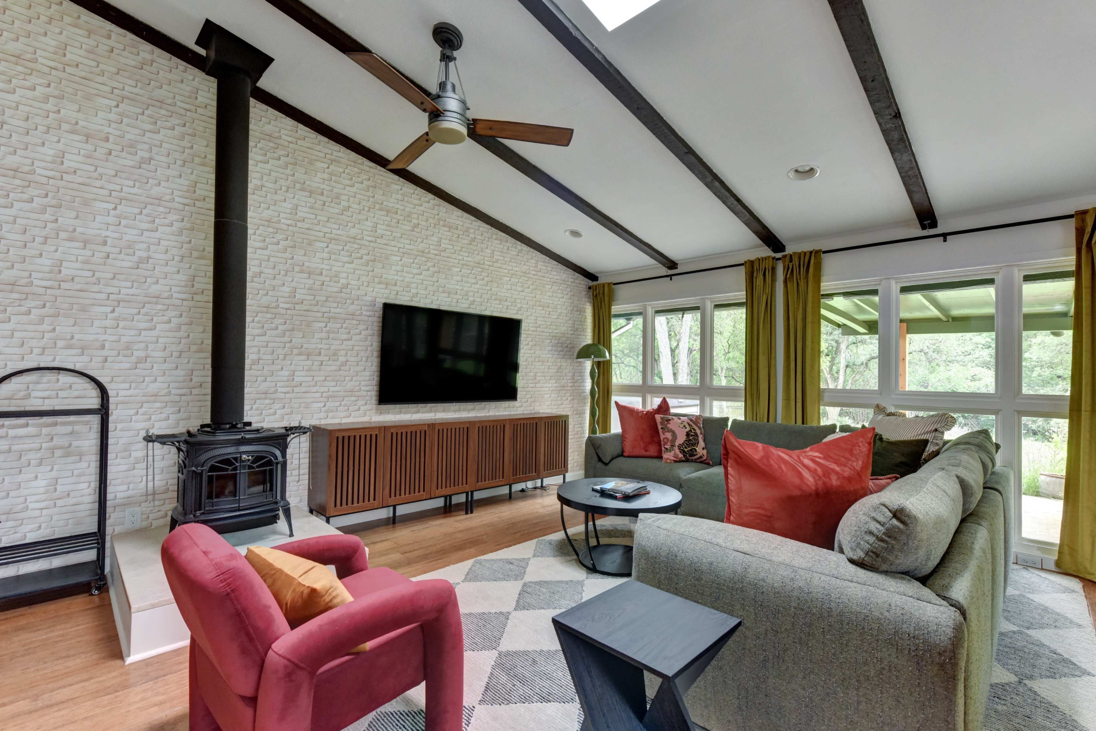 The living room features a gray sectional sofa, a burgundy armchair, a wood-burning stove, and large windows overlooking a green outdoor area.