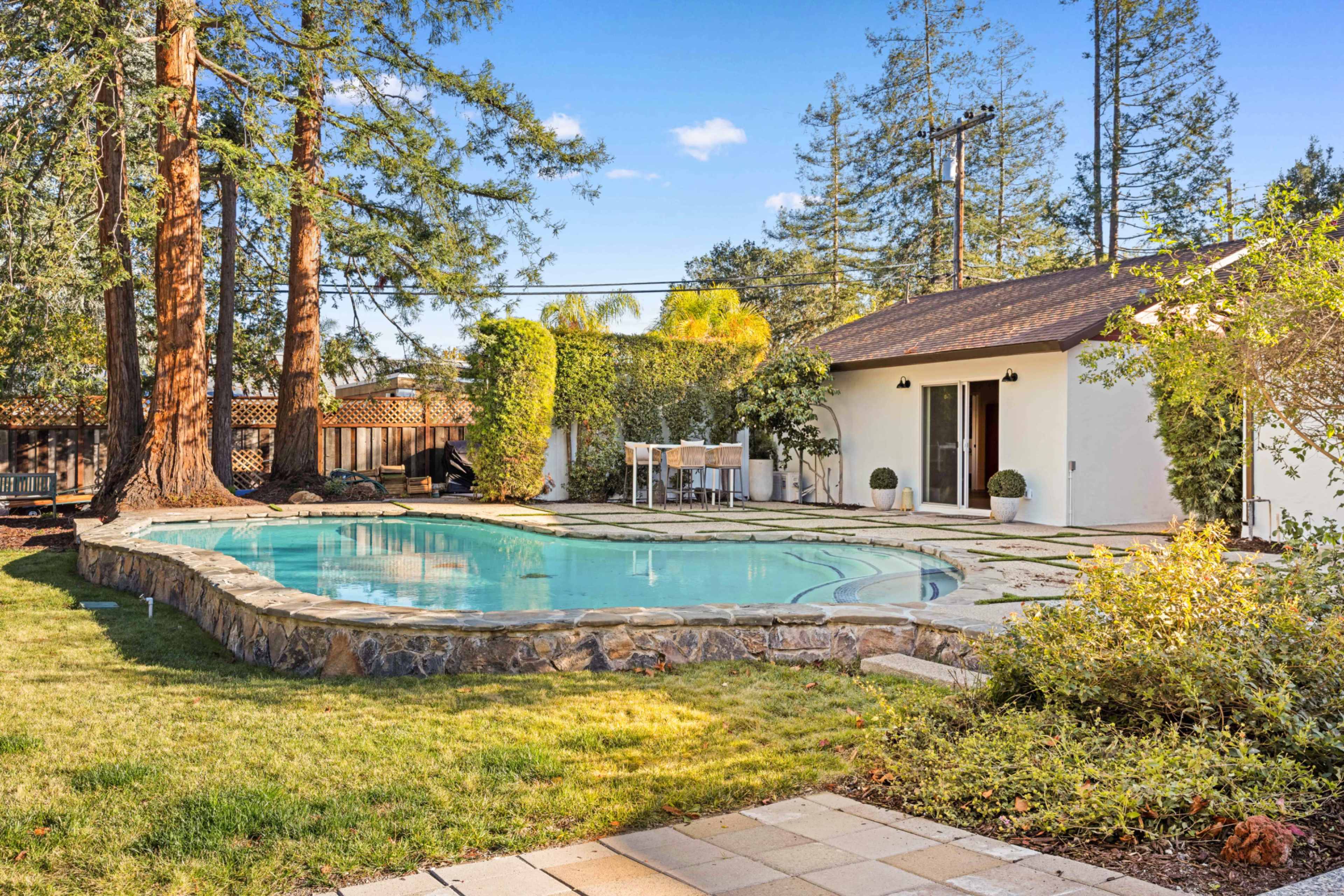 The image shows a residential backyard featuring a pool surrounded by a stone patio, lawn, and several trees, with a house visible in the background.