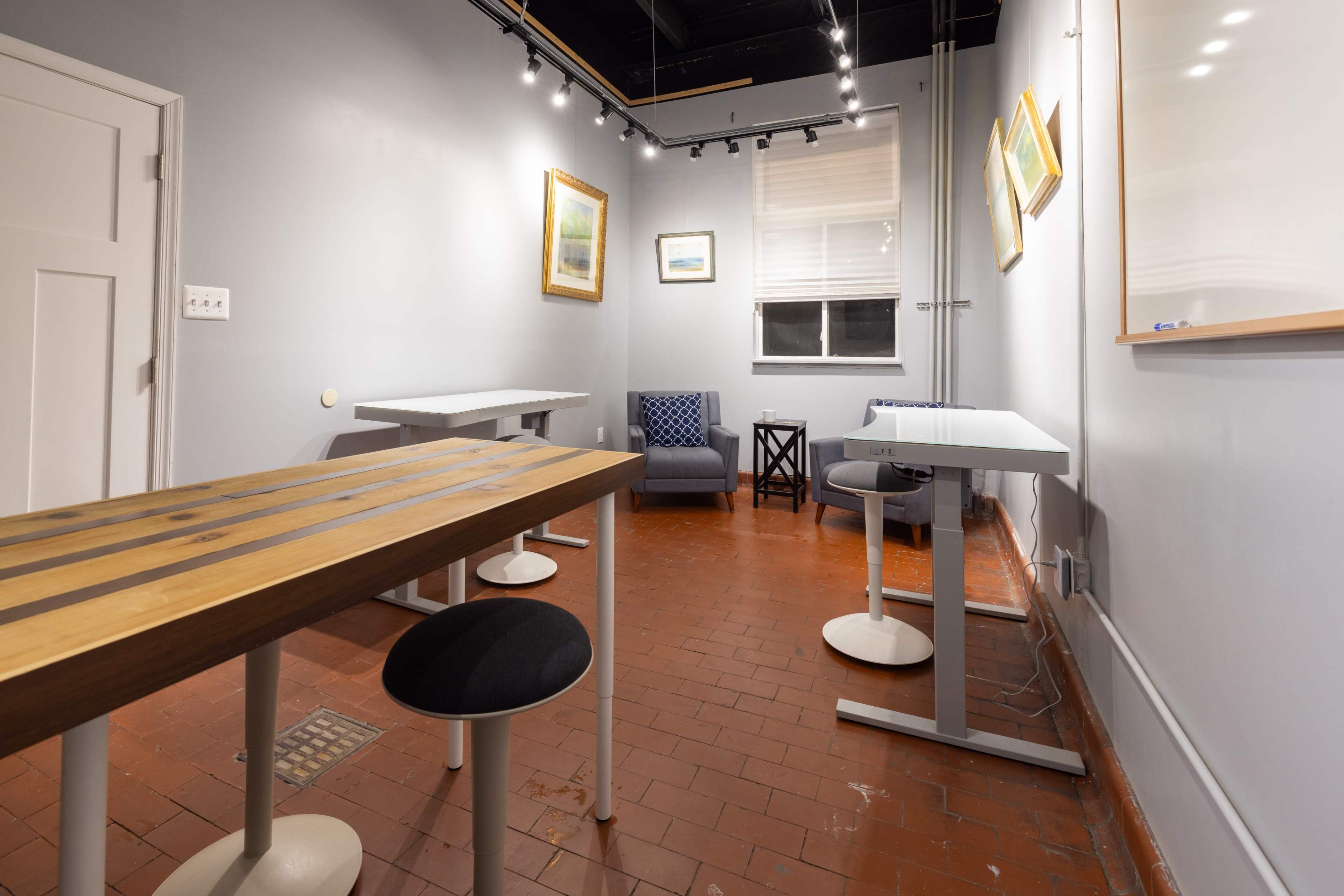 A small, minimalist room with two tables, several stools, and a chair, along with framed pictures on the walls and a window providing natural light.