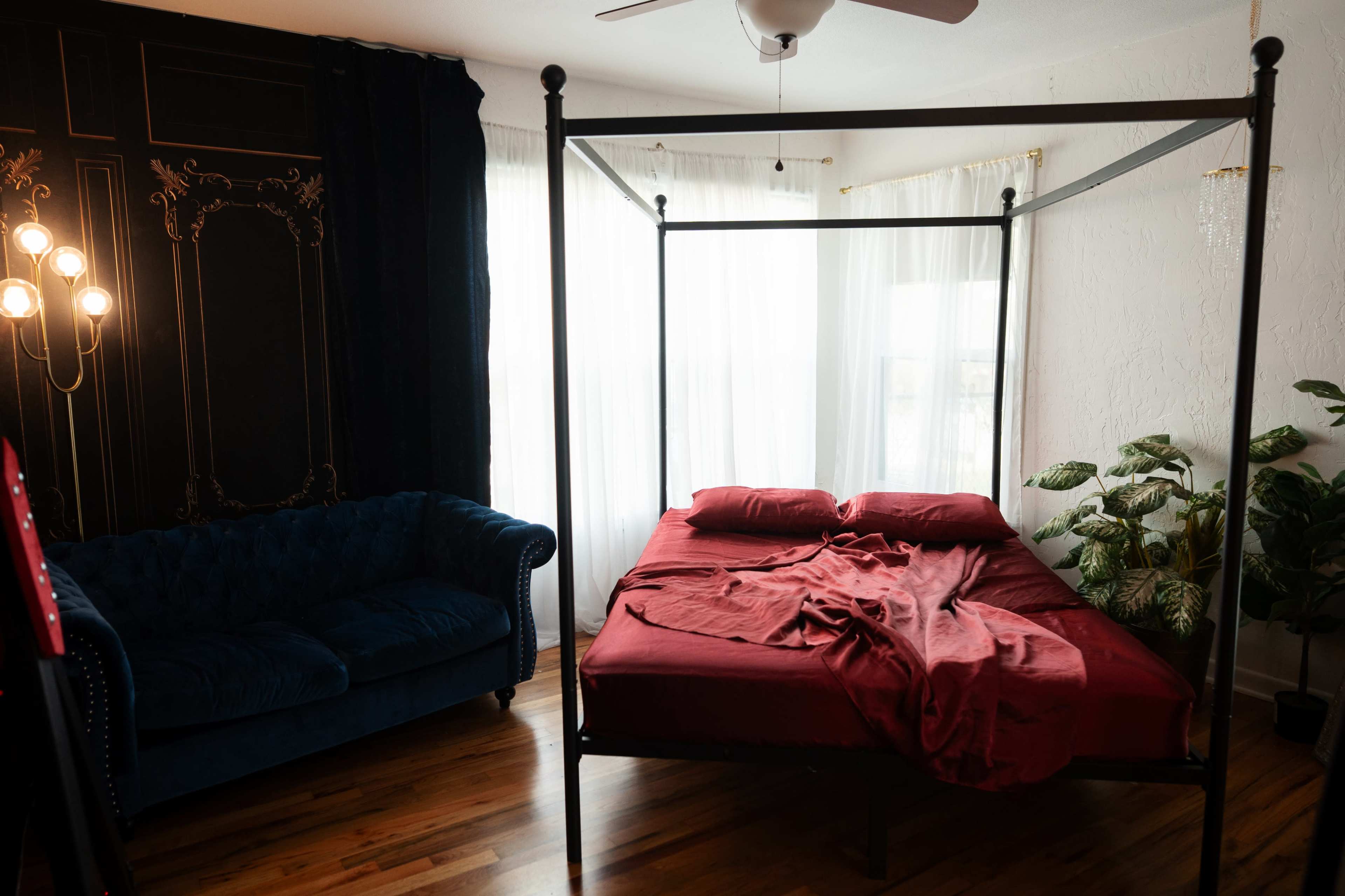 The image shows a bedroom with a four-poster bed covered in a red blanket, a dark blue couch, and large windows allowing natural light.