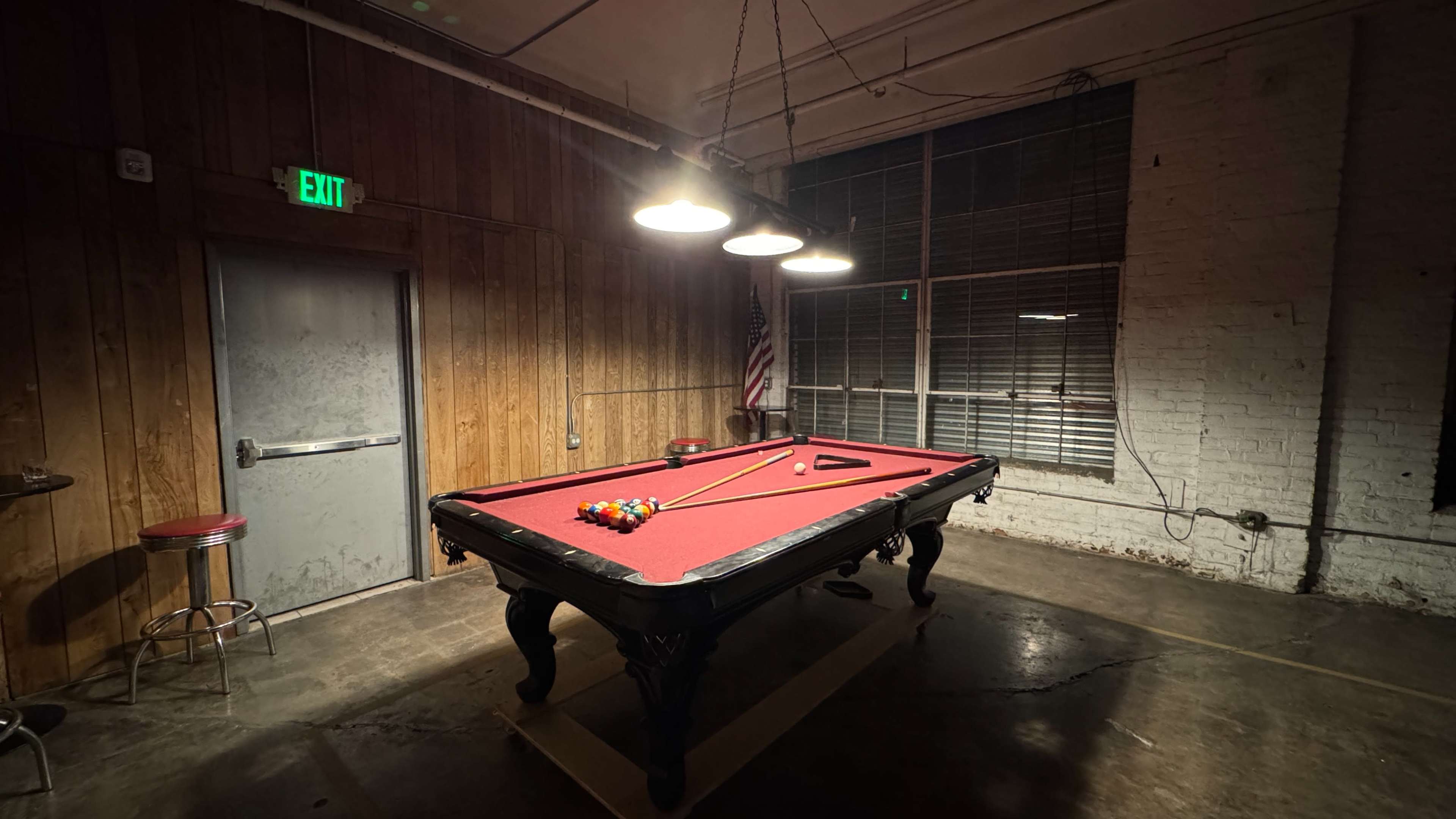 A red felt pool table is set up in a dimly lit room with wooden walls and a large window, featuring three overhead lights and an American flag in the background.