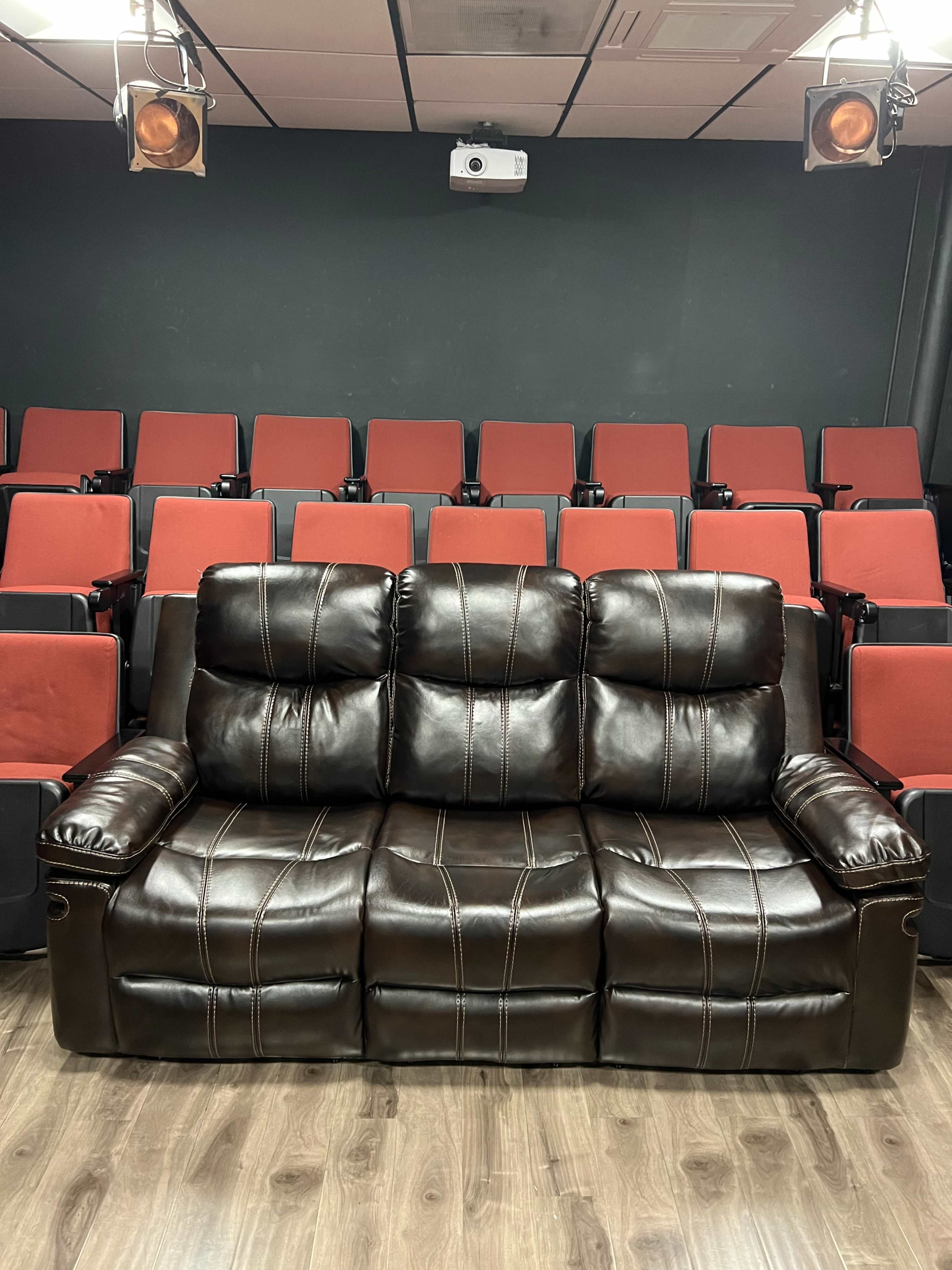 A brown leather couch with three seats is positioned in front of a row of red theater-style seats in a screening room.