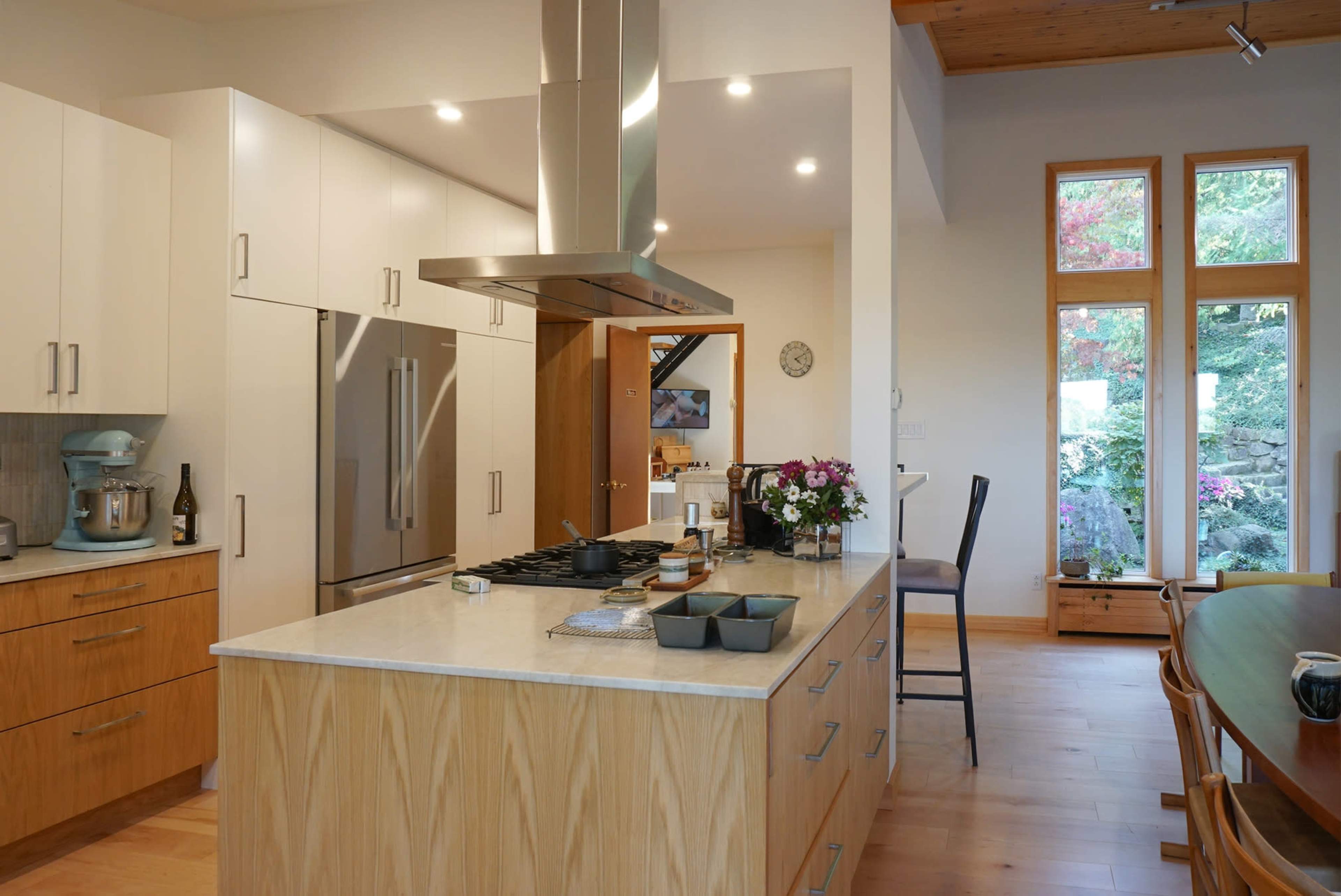 A modern kitchen features a large island with a countertop, stainless steel appliances, and a wooden dining table beside large windows.