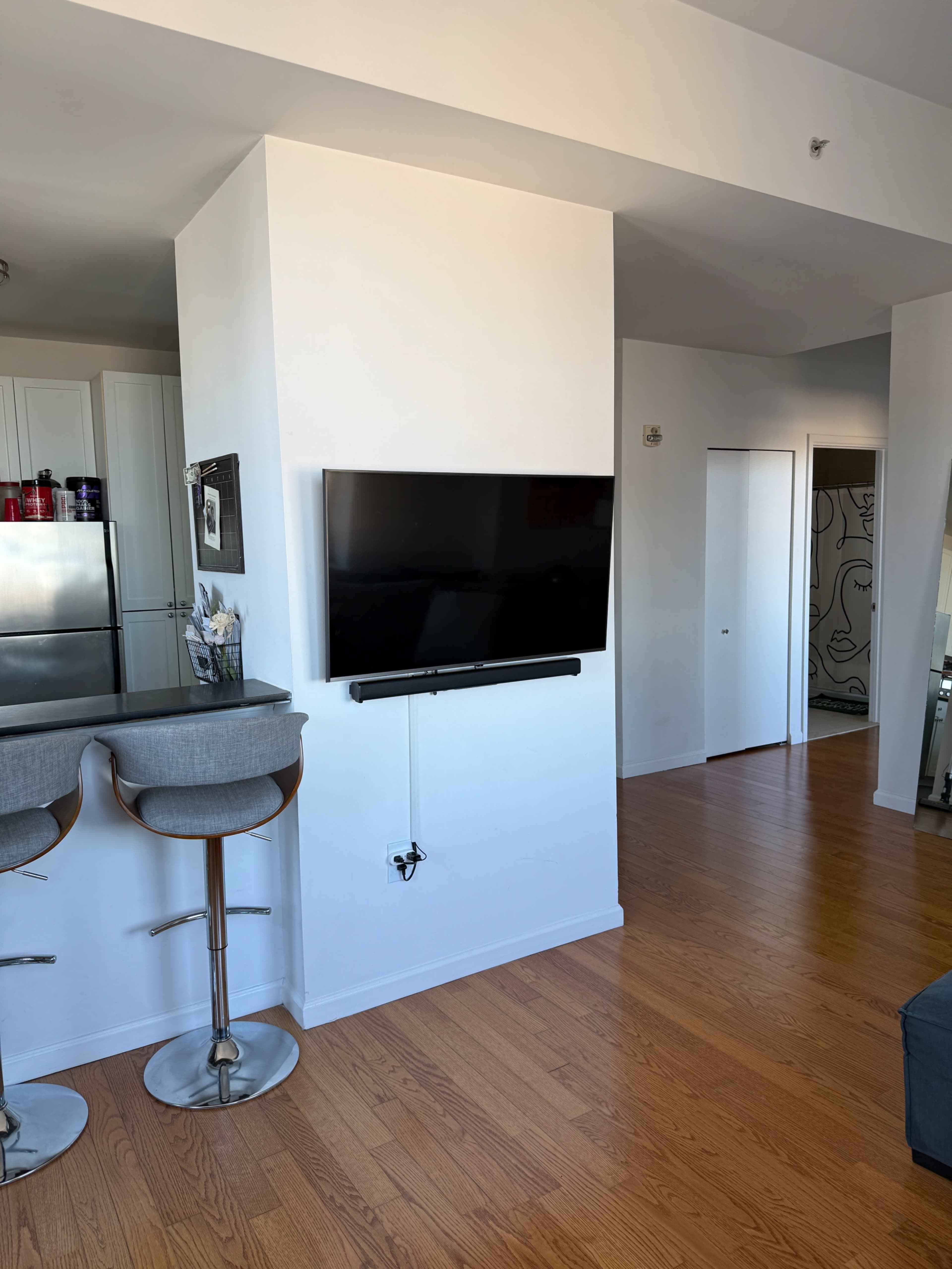 The image shows a modern living space with a mounted flat-screen TV on a white wall, two bar stools at a kitchen counter, and an open doorway leading to another room.