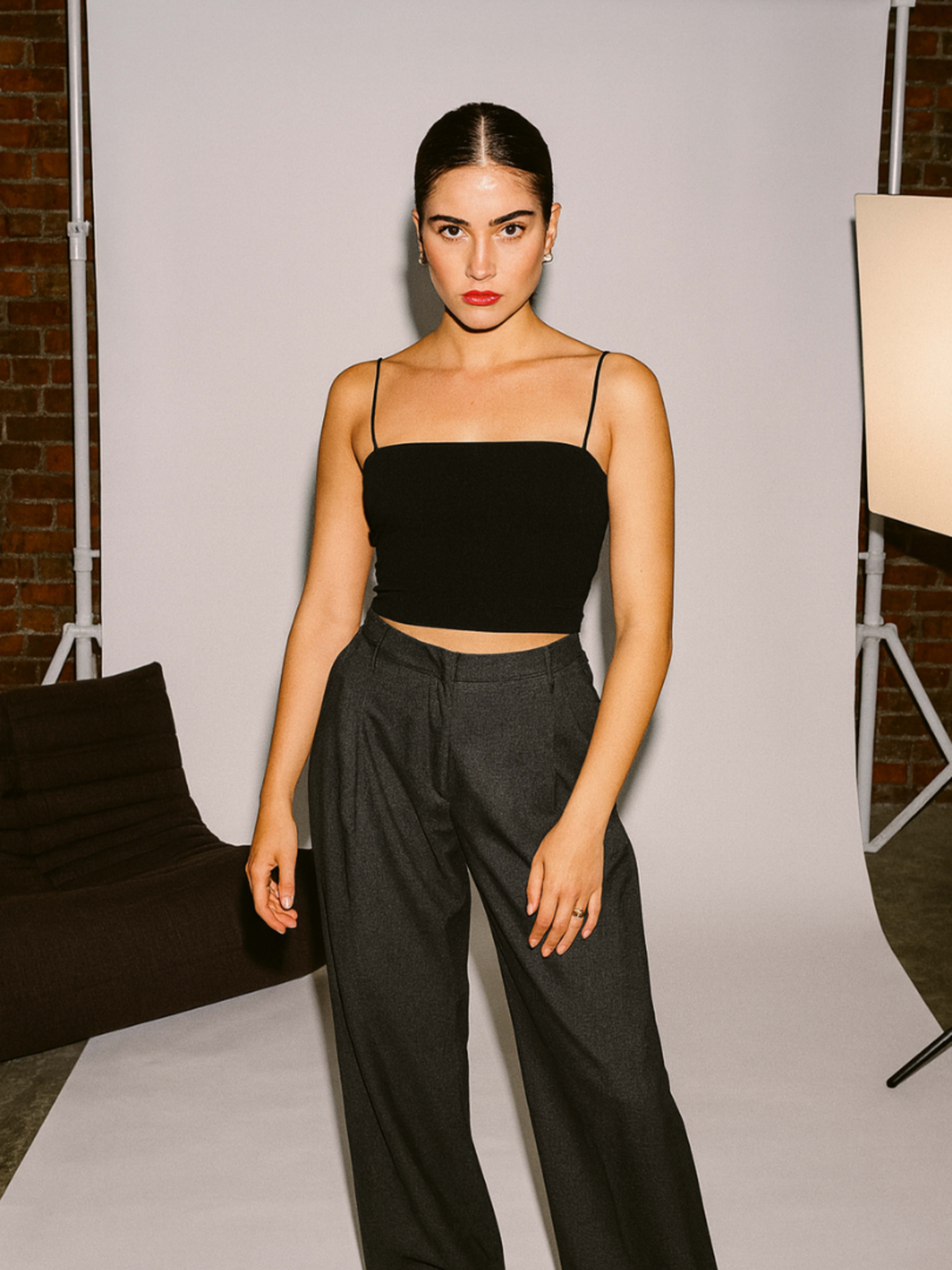 A woman stands confidently in a black crop top and gray trousers against a neutral backdrop in a studio setting.