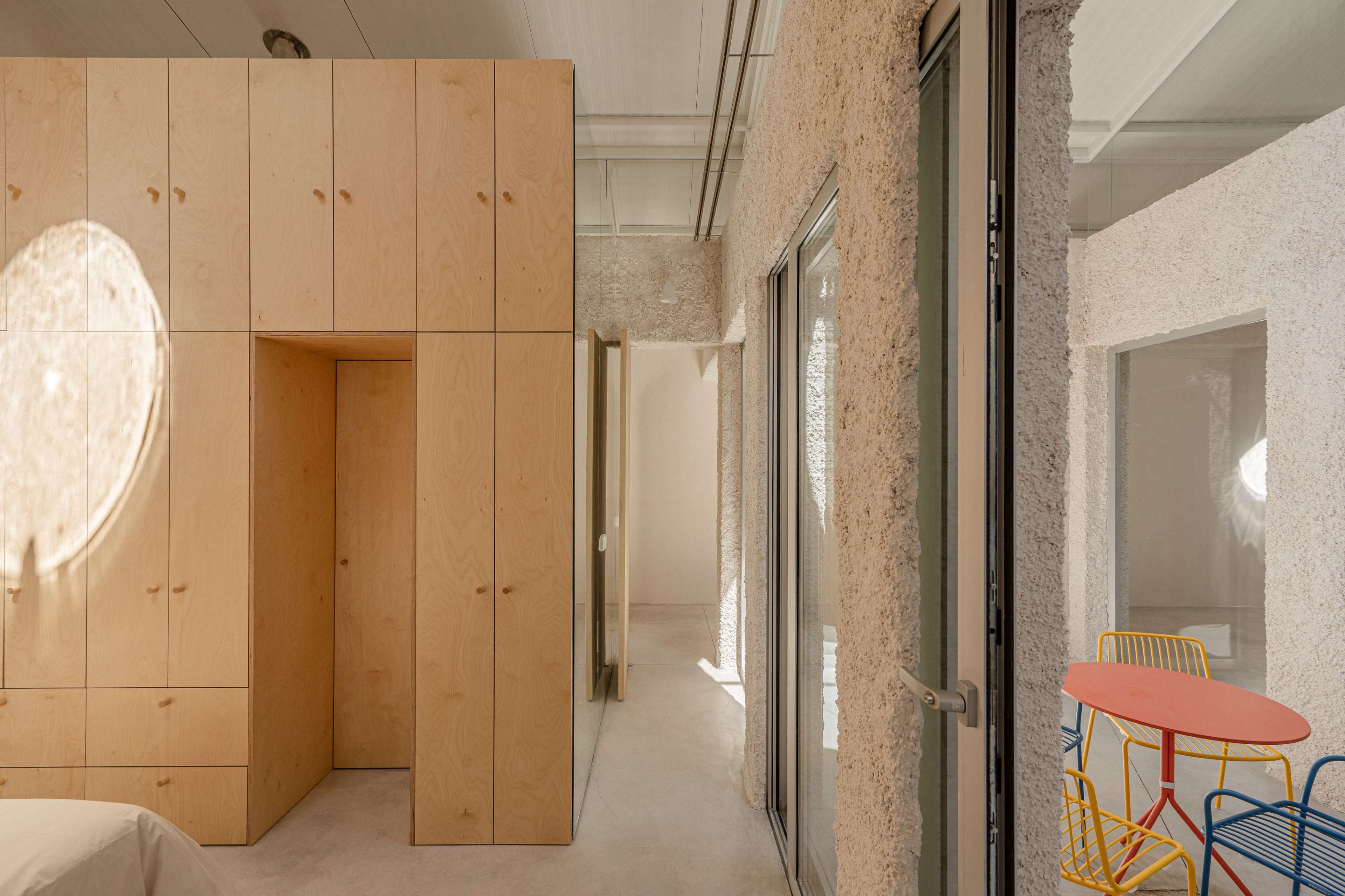 The image shows a modern interior space featuring light-colored walls, a wooden wardrobe unit, and colorful furniture, including a round table and chairs.